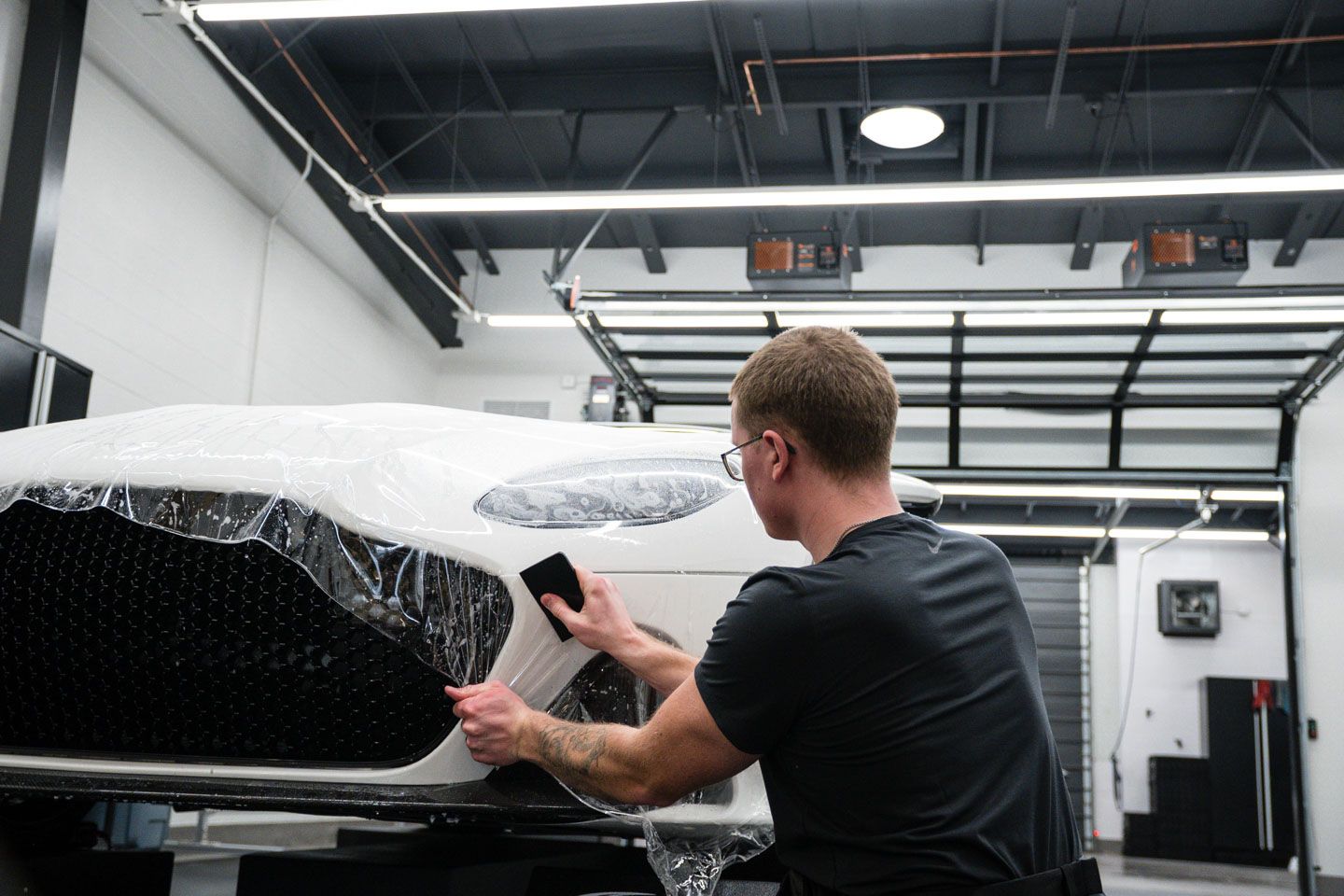 A person applying protective film to a white car's front bumper in a garage. They use a squeegee to smooth the film.