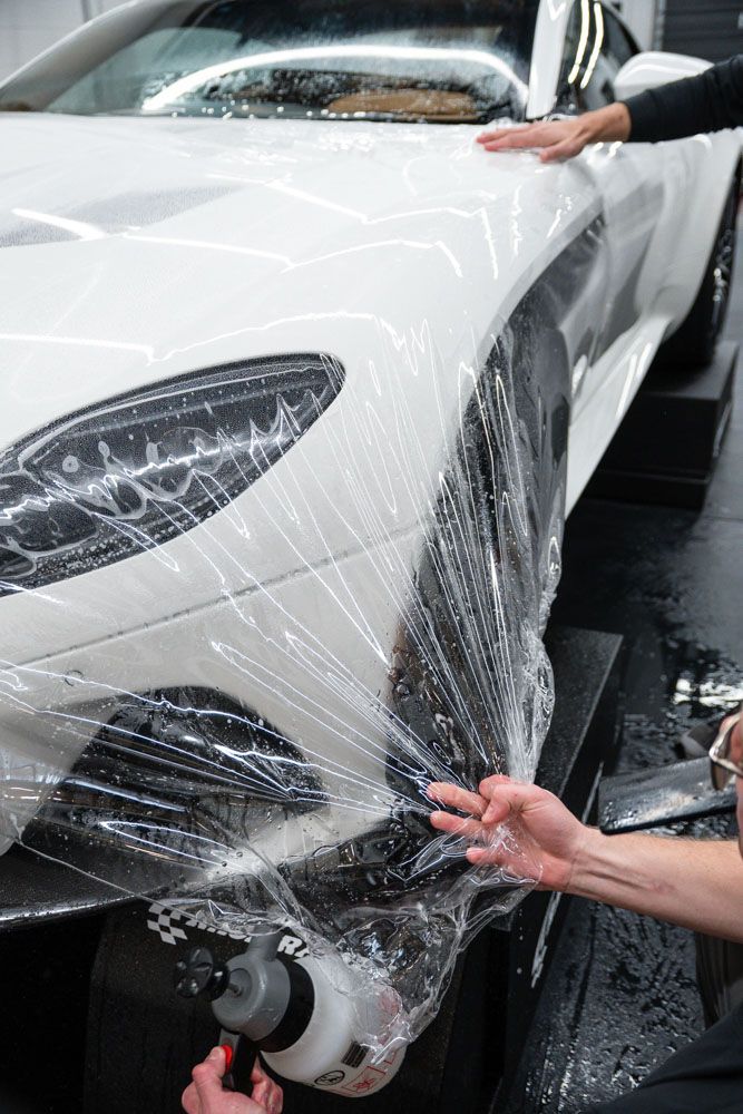 Workers applying clear protective film to the front of a white car, spraying it with a bottle in a garage setting.