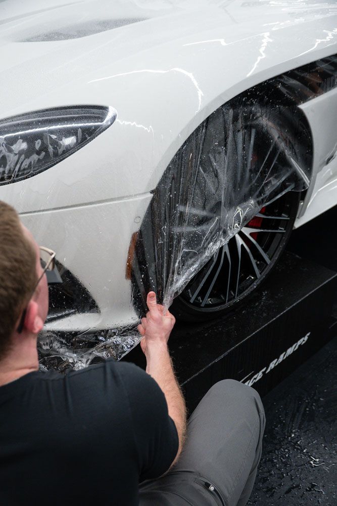 Man applying clear protective film to the front fender of a white car. He is kneeling, working on the wheel well.