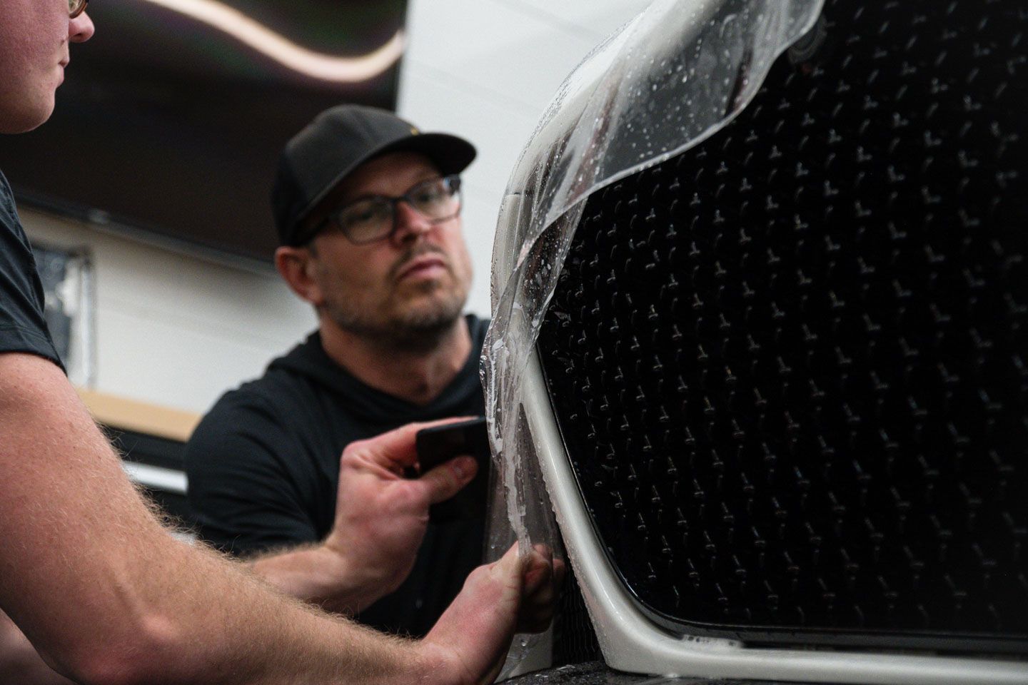Person in black gloves applying sealant to a black car's hood in a well-lit garage.