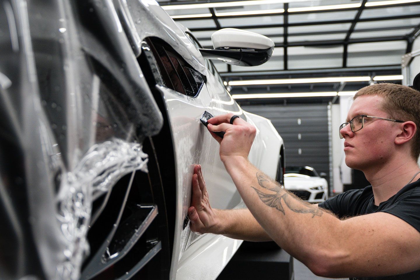 A man in glasses applies protective film to a white car door in a garage. He uses a tool and his hands.