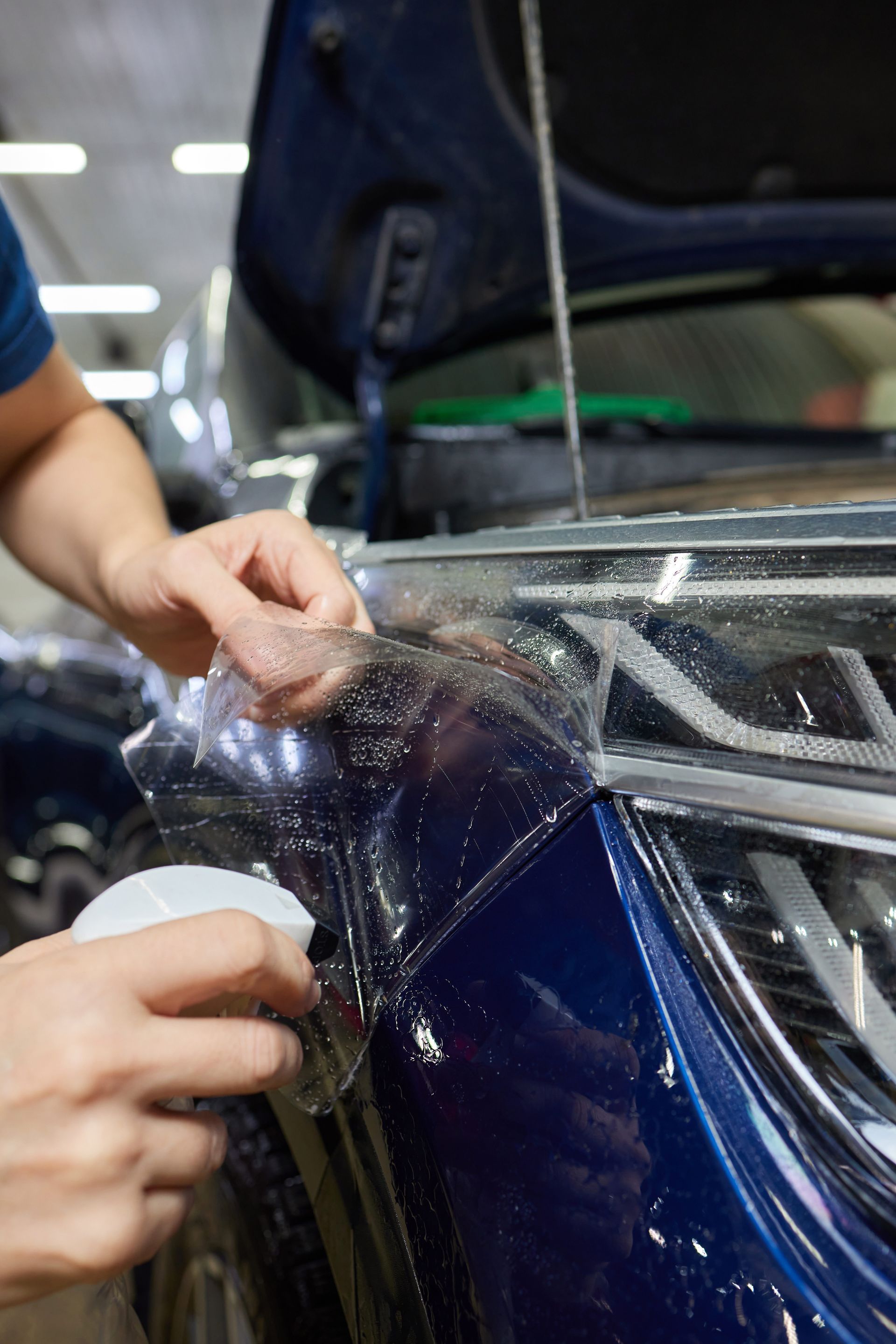 Person applying clear protective film to the front of a blue car. The car's hood is open.