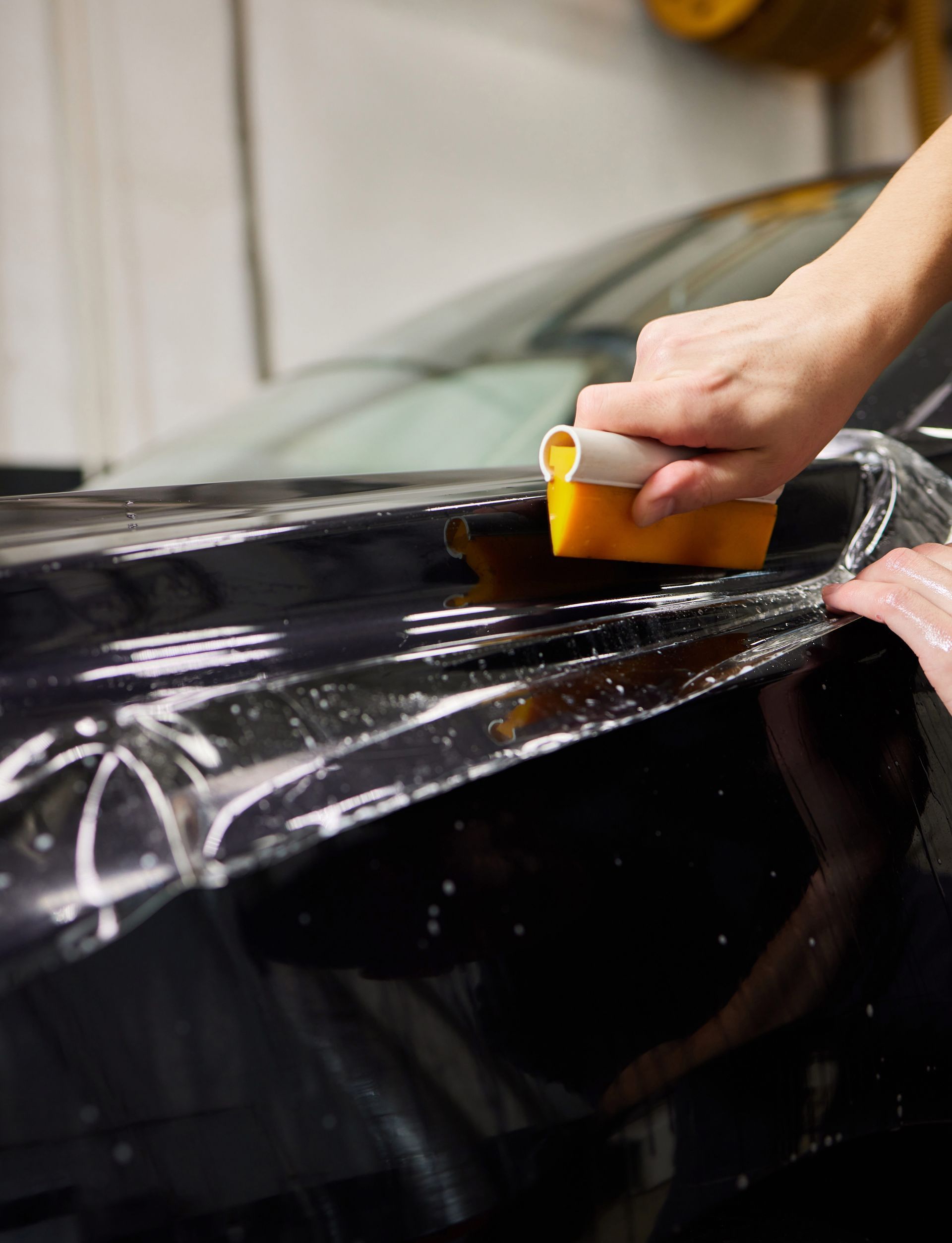 Person applying clear protective film to the black side panel of a car, smoothing it with a squeegee in a workshop.