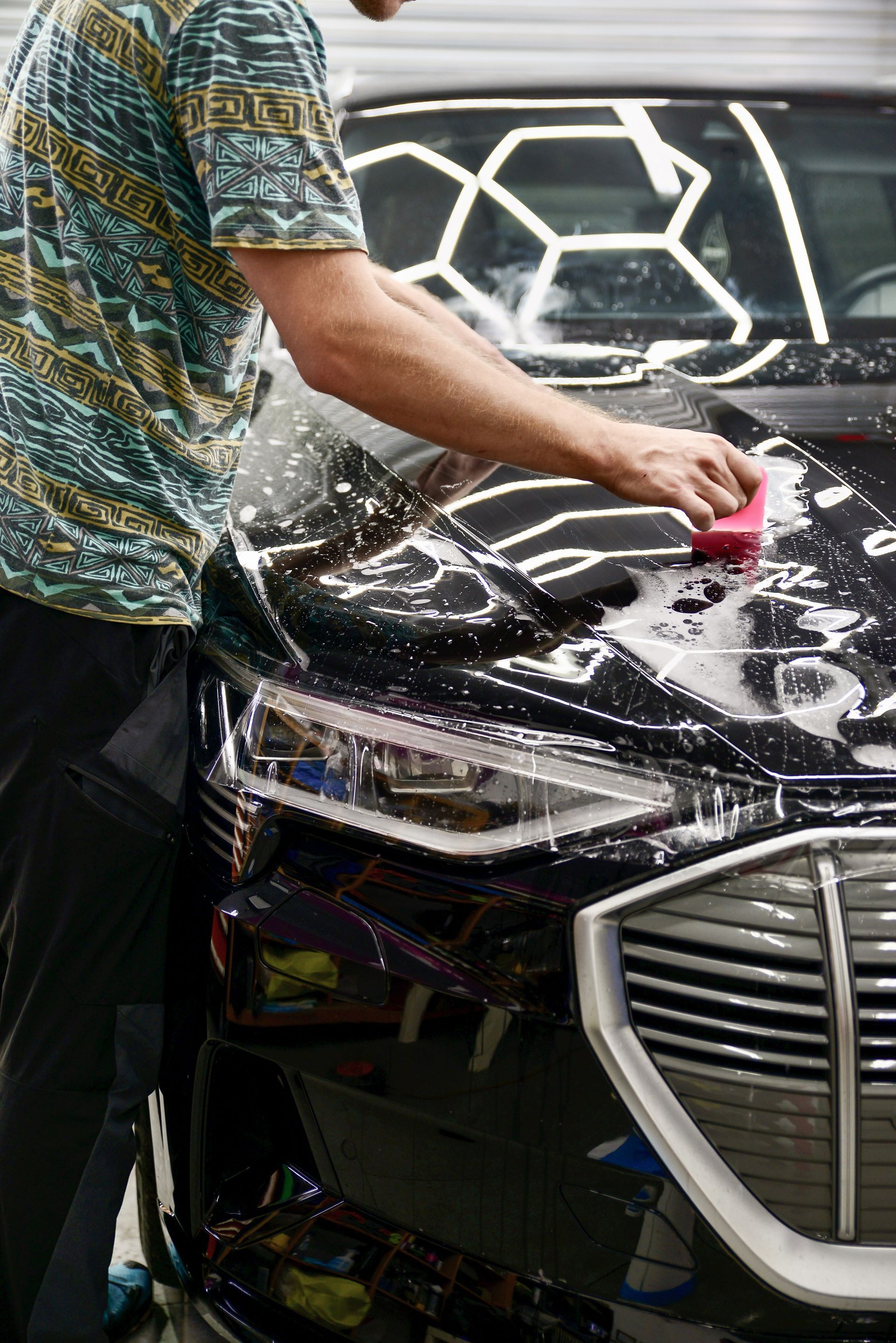 A person washes a black car, scrubbing its hood with a red sponge. The car is in a well-lit space.