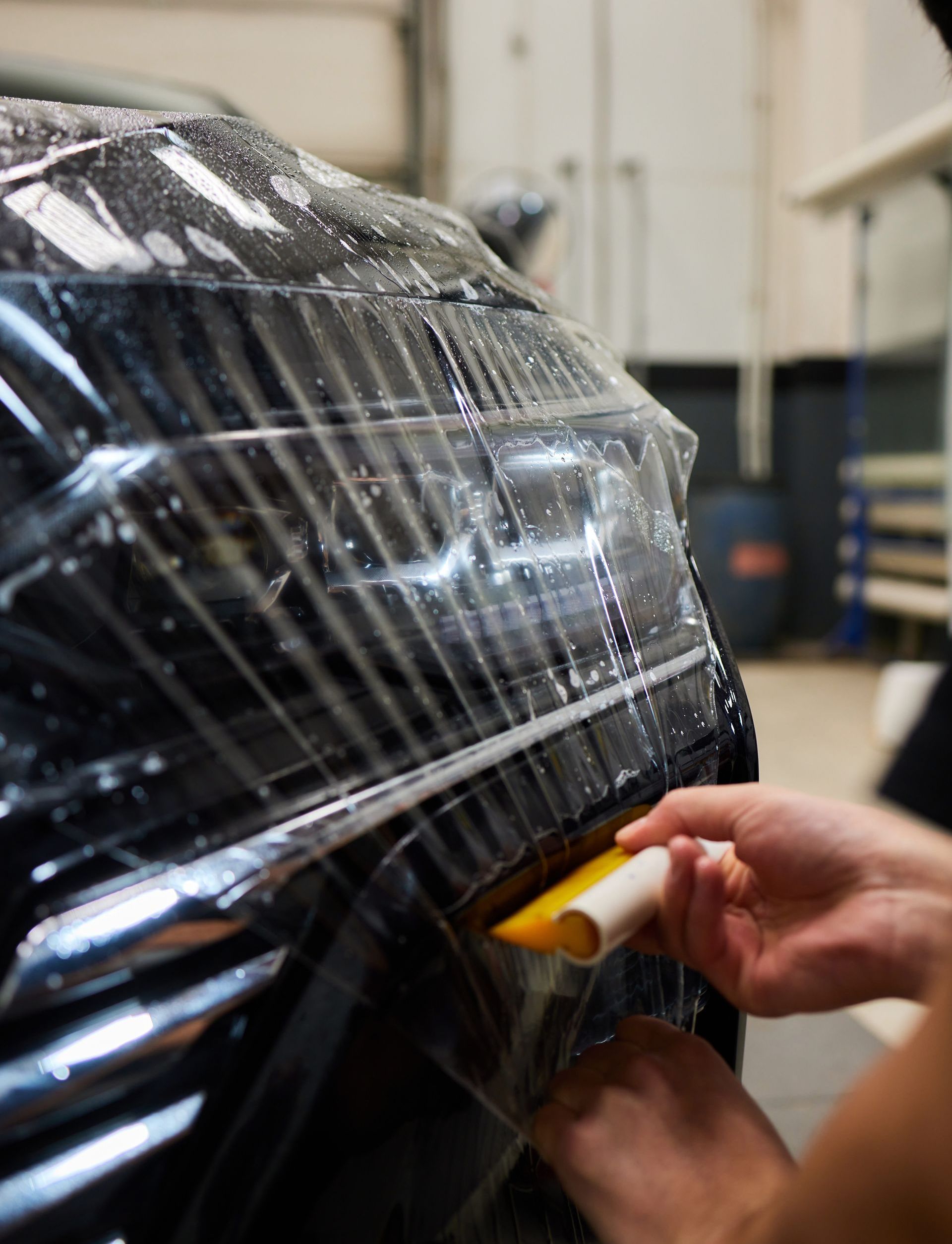 Person applying clear protective film to a black car, using a squeegee. Inside a well-lit workshop.