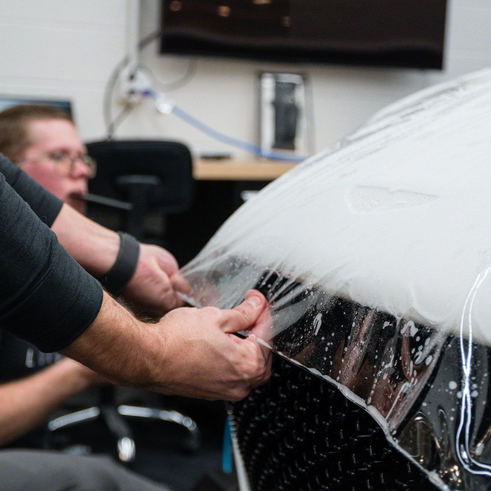 Dark green sports car's hood, with vents, reflected in a showroom window, on a gray floor.