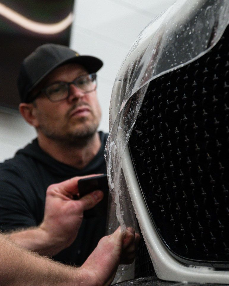 Person in black gloves applying sealant to a black car's hood in a well-lit garage.