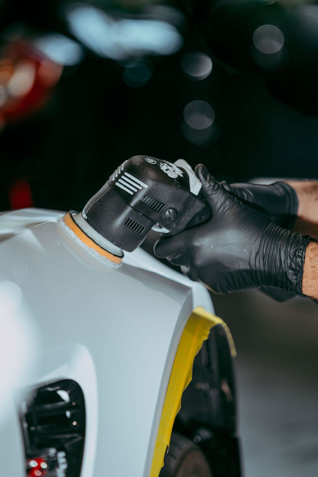 A person wearing black gloves polishes a car's white fender with a black electric buffer, in a dim setting.