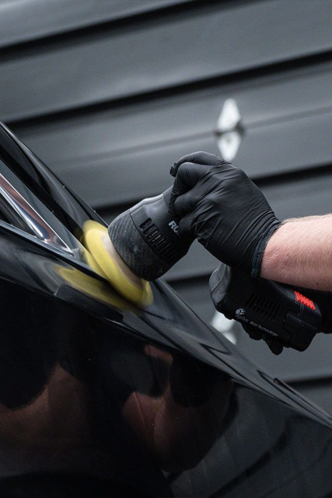 Person wearing black gloves polishing a black car with a buffing tool.