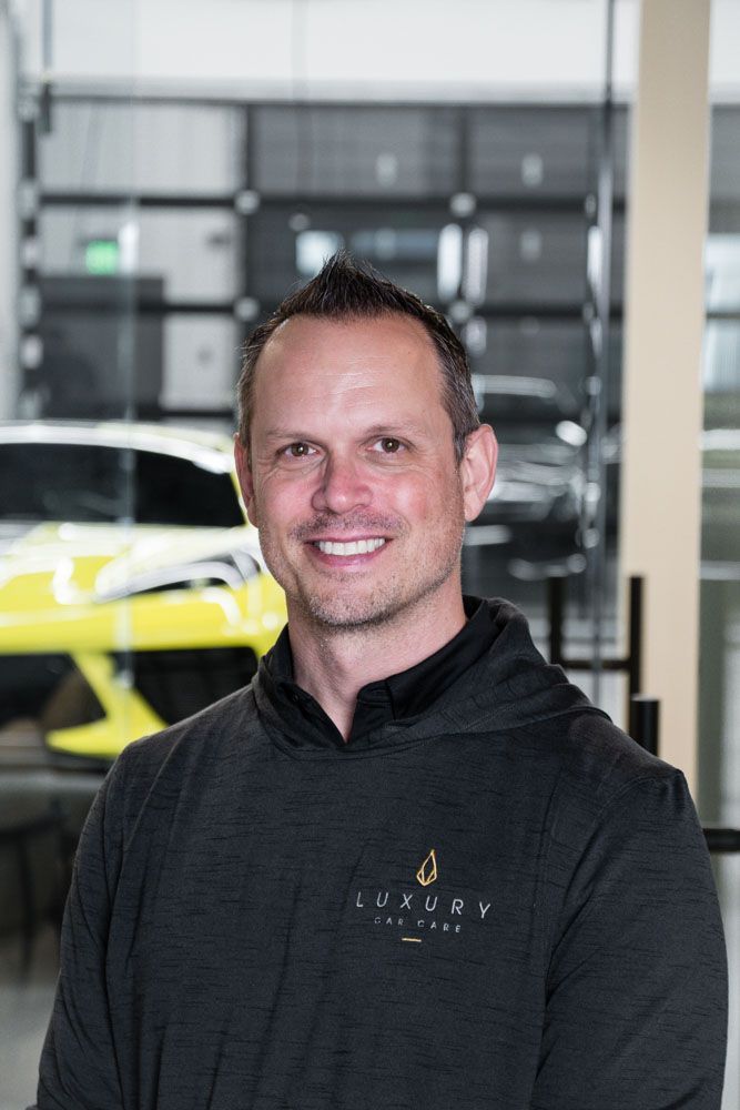Man in a dark hoodie smiles in front of a car showroom, with a bright yellow car in the background.