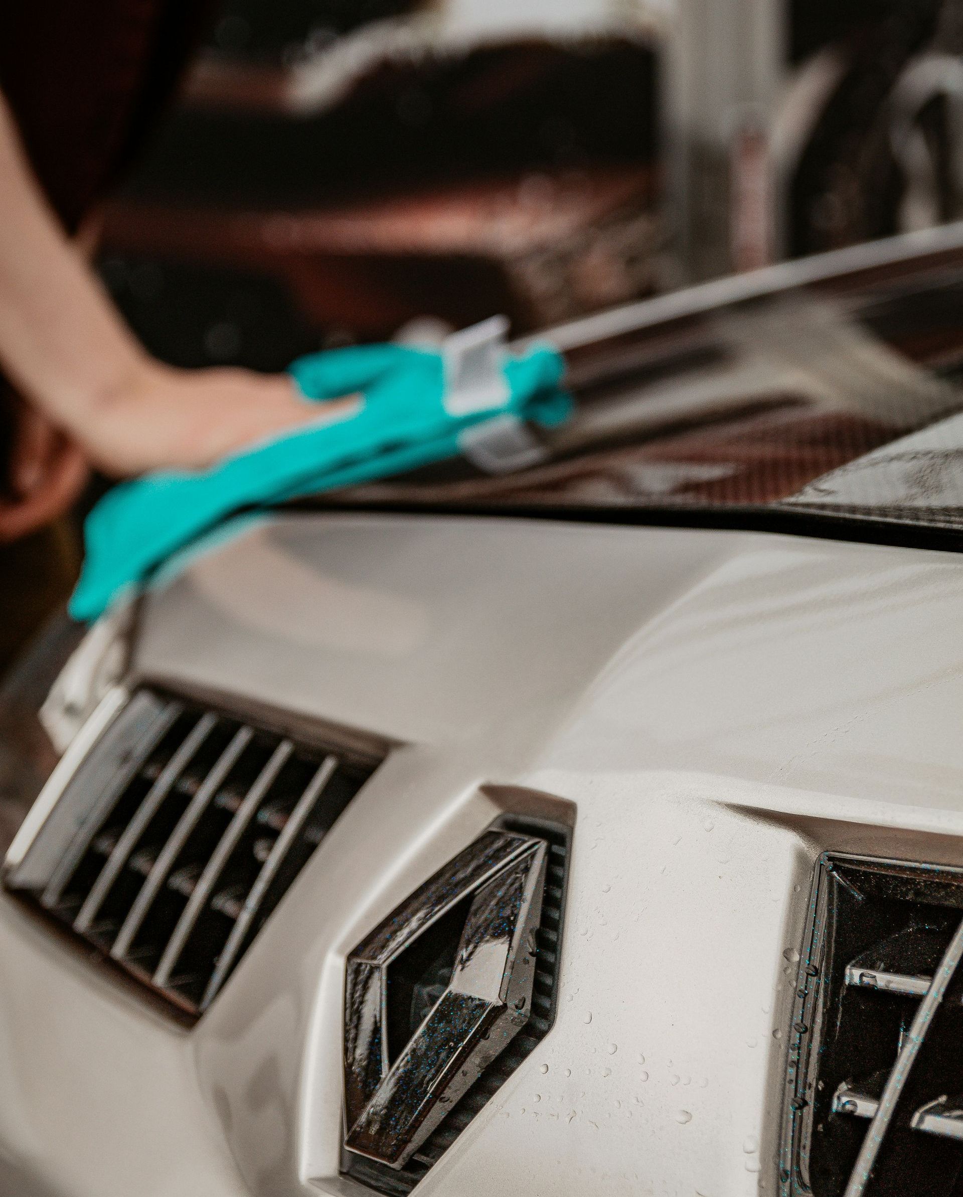A person cleaning a white car's hood with a turquoise cloth. Focus on the car's front, showing a grill and headlight.