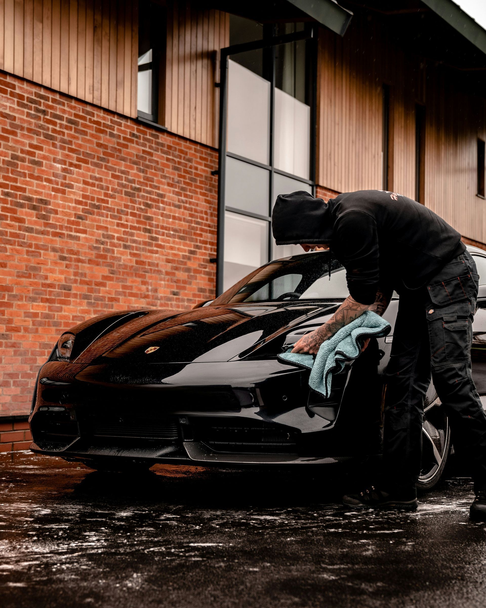 A person in black clothing wipes down a black car in front of a brick and wood building.
