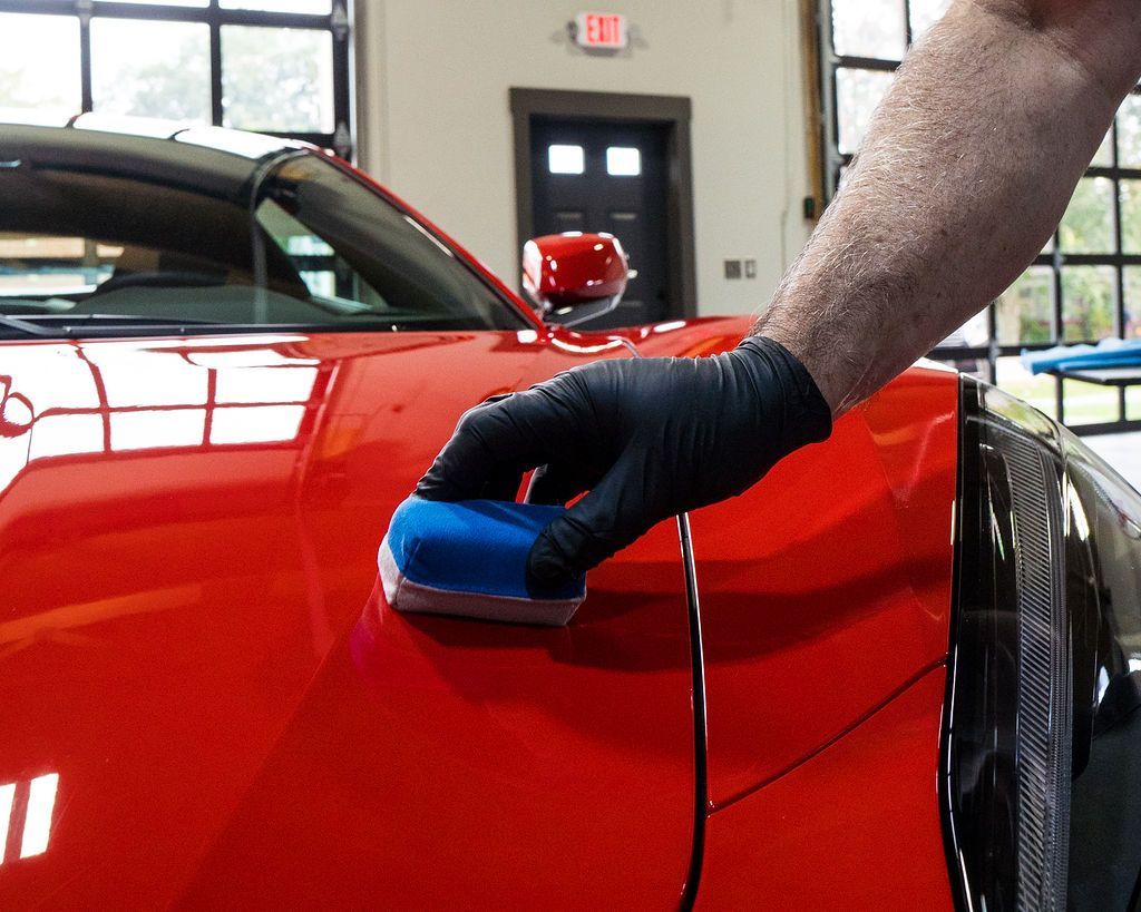 A person wearing a black glove applies a blue foam applicator to the red car's hood. The car is inside a garage.