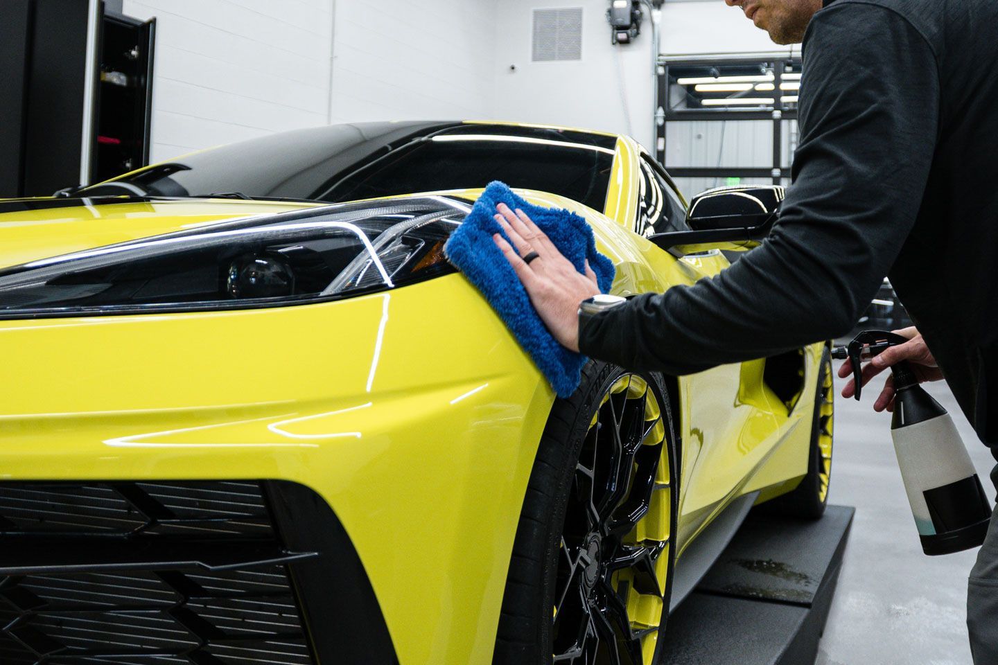 A person wiping a yellow sports car with a blue cloth. They are standing in a garage.