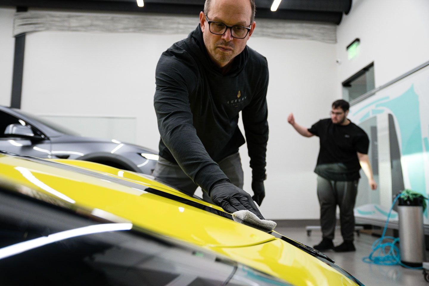 Man in glasses, wearing gloves, wiping a yellow car hood in a shop, with another man gesturing in the background.