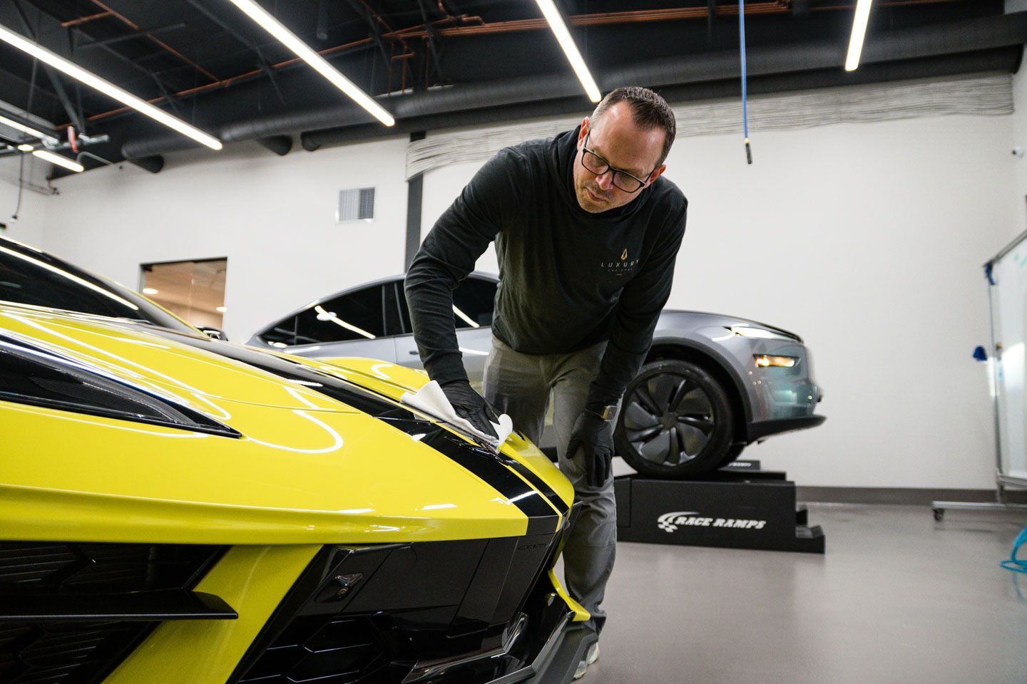 Man inspecting the front of a yellow sports car in a garage, a gray car in the background.