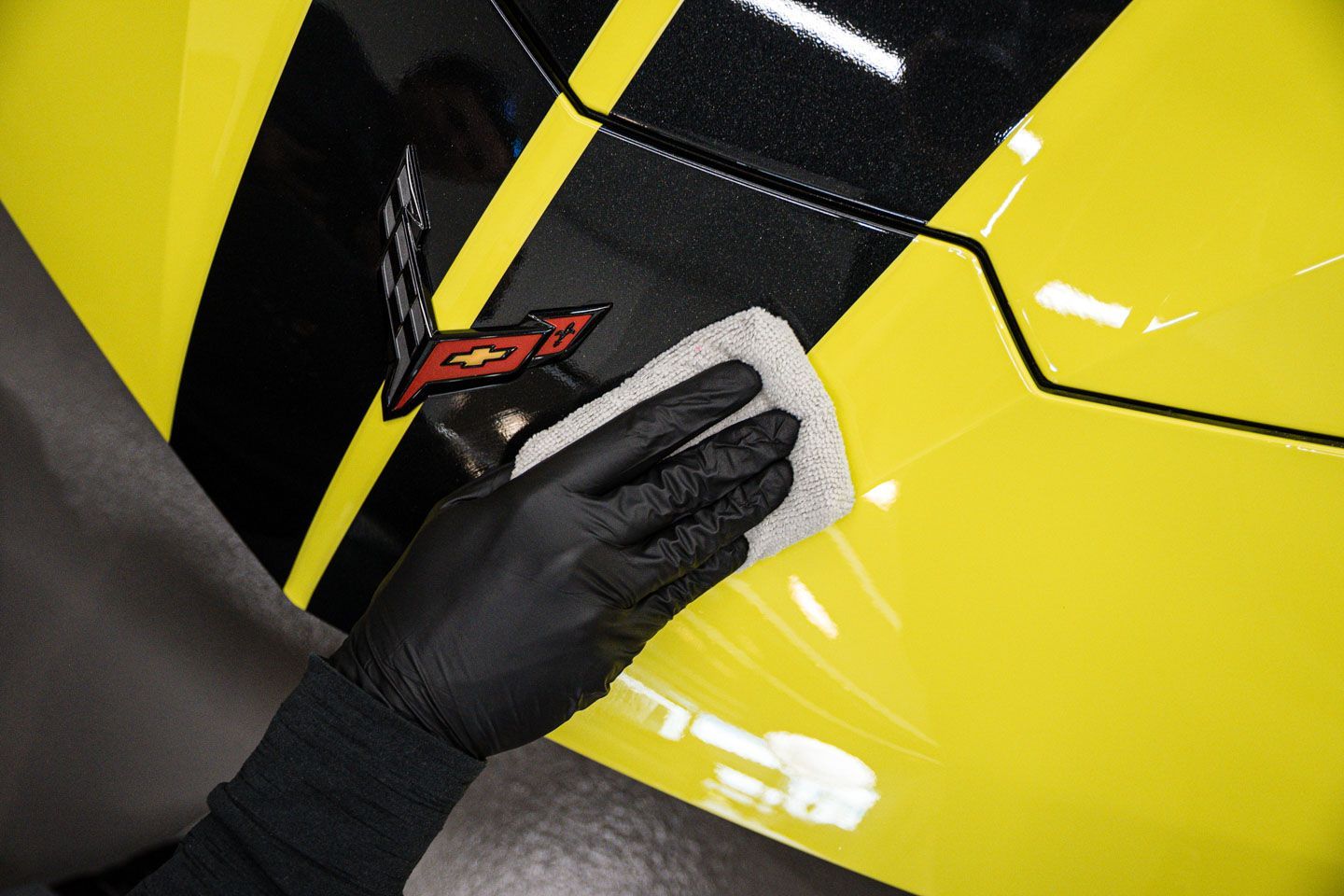 A gloved hand wipes a yellow car with a white cloth, near a black stripe and a red logo.
