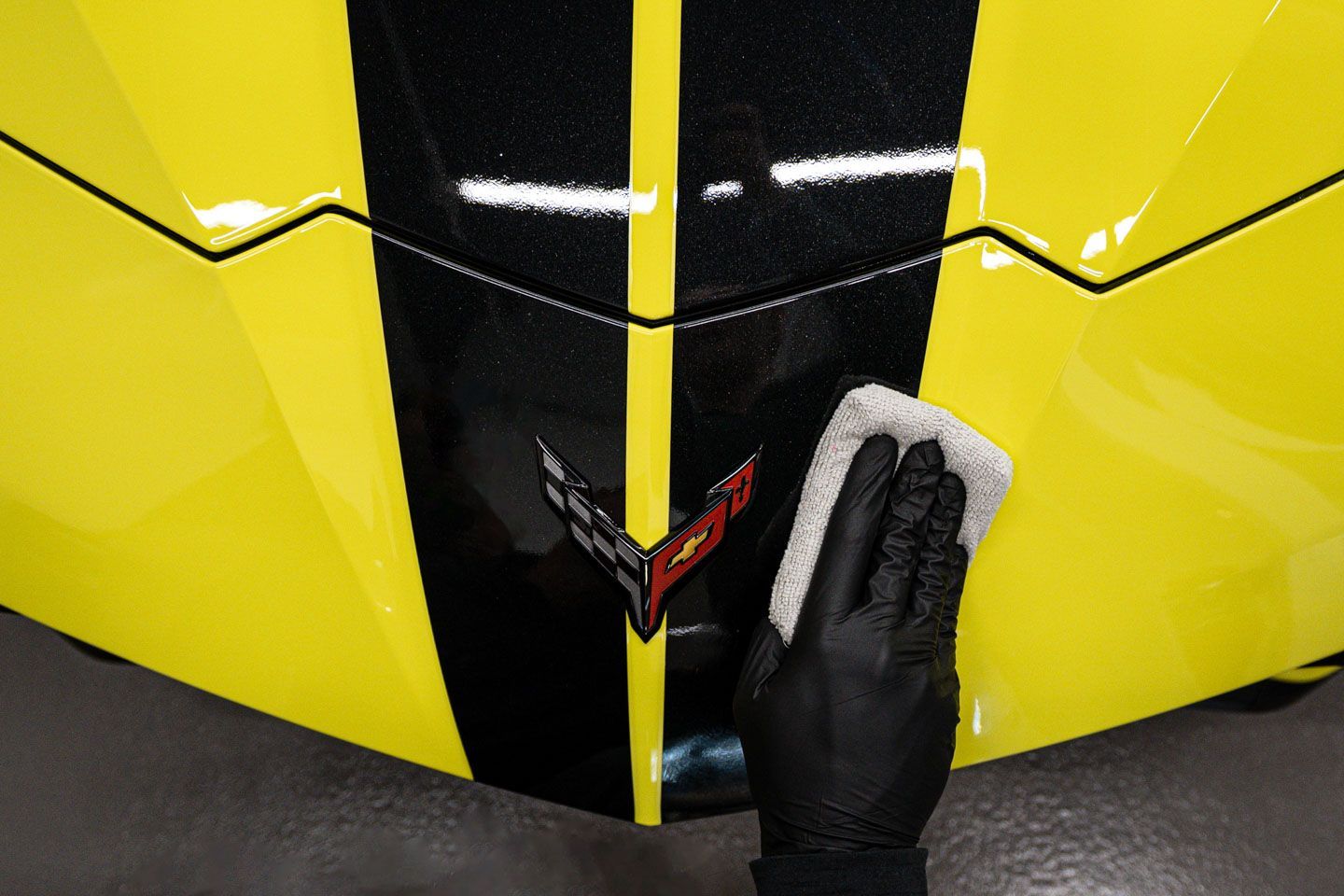 A gloved hand wipes a white cloth across the black stripe on a yellow Corvette hood, near its emblem.