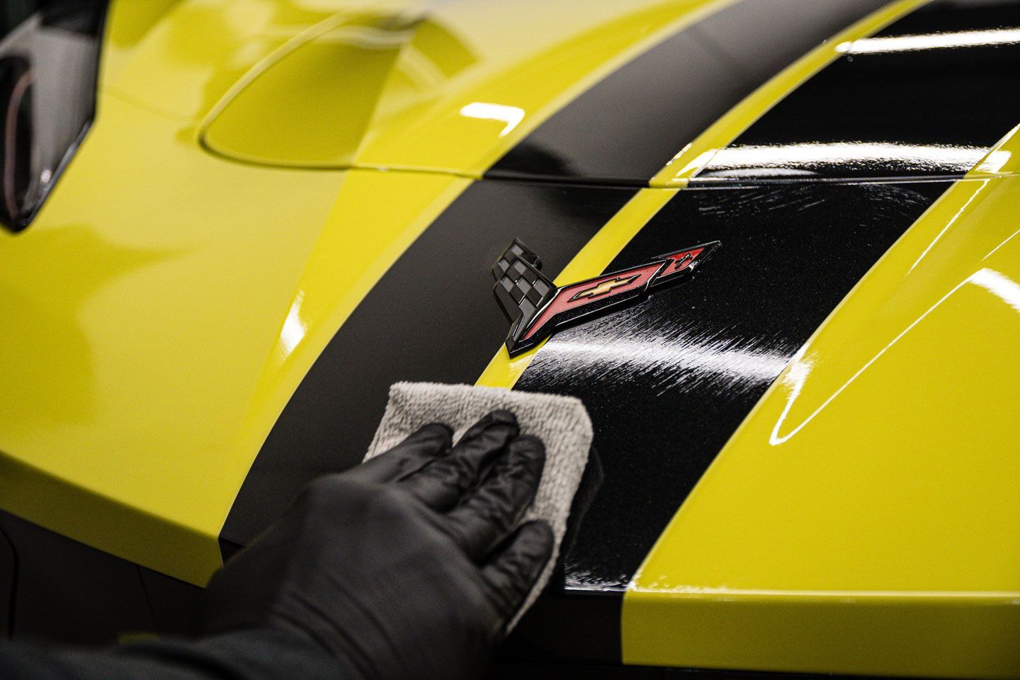 Hand cleaning a black stripe with a cloth on a yellow Corvette's hood, near the emblem.
