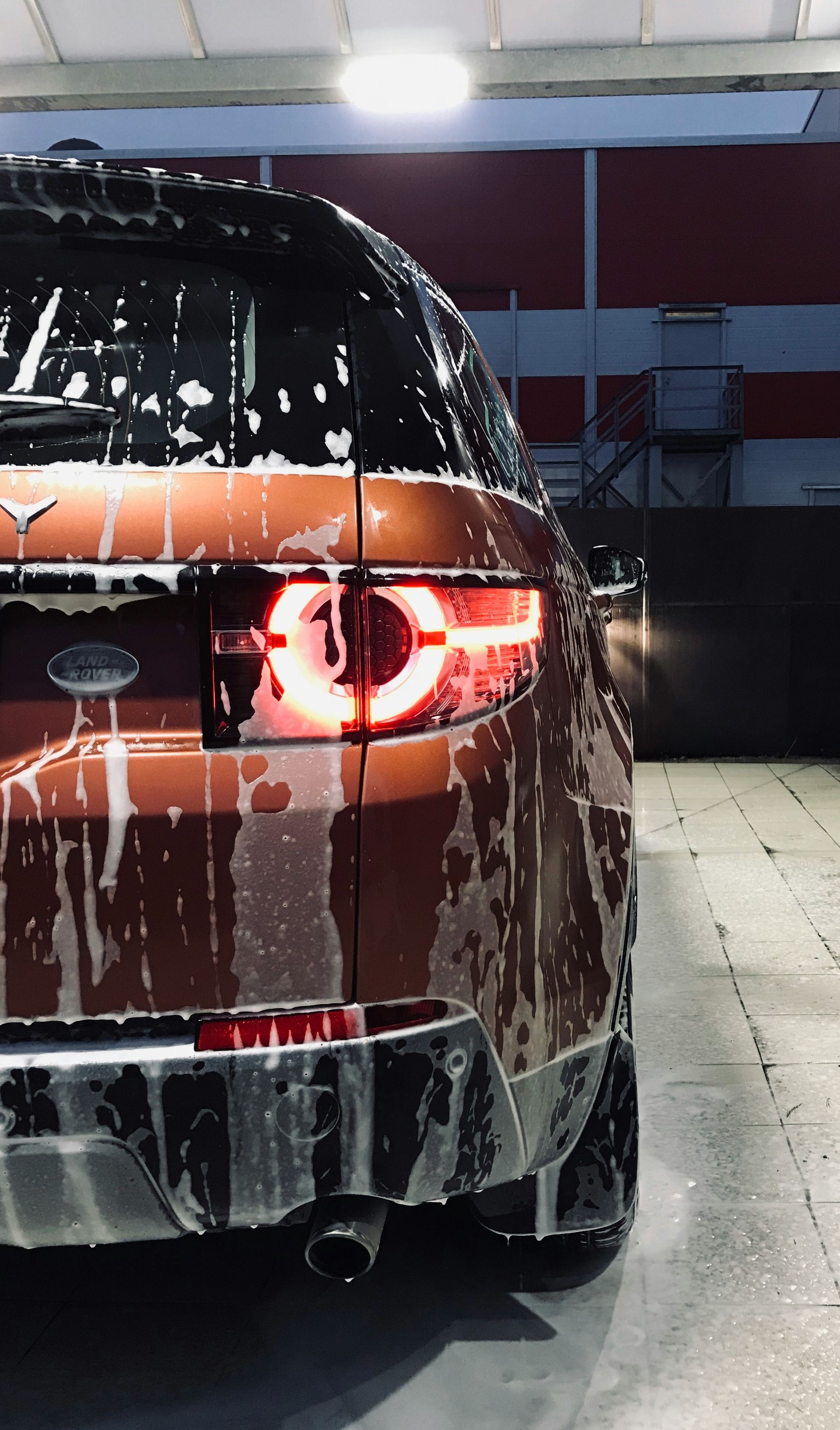 Car covered in soap suds at a car wash, rear view showing taillight and exhaust.