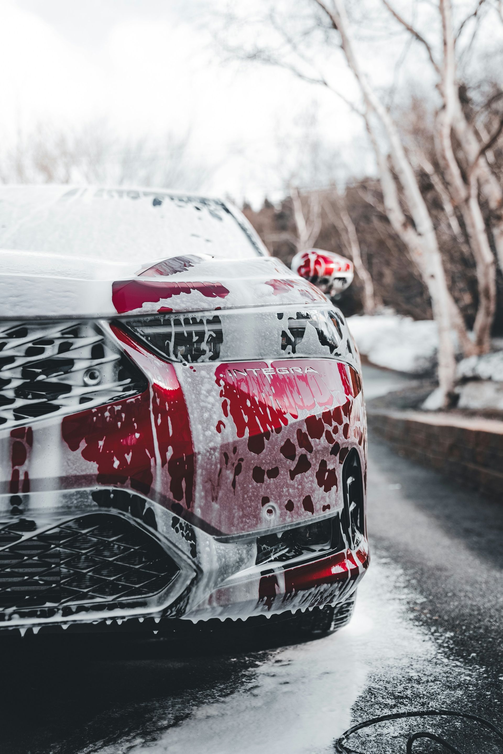 A red car being washed with foamy soap, outside on a paved road with trees and a snowy bank.