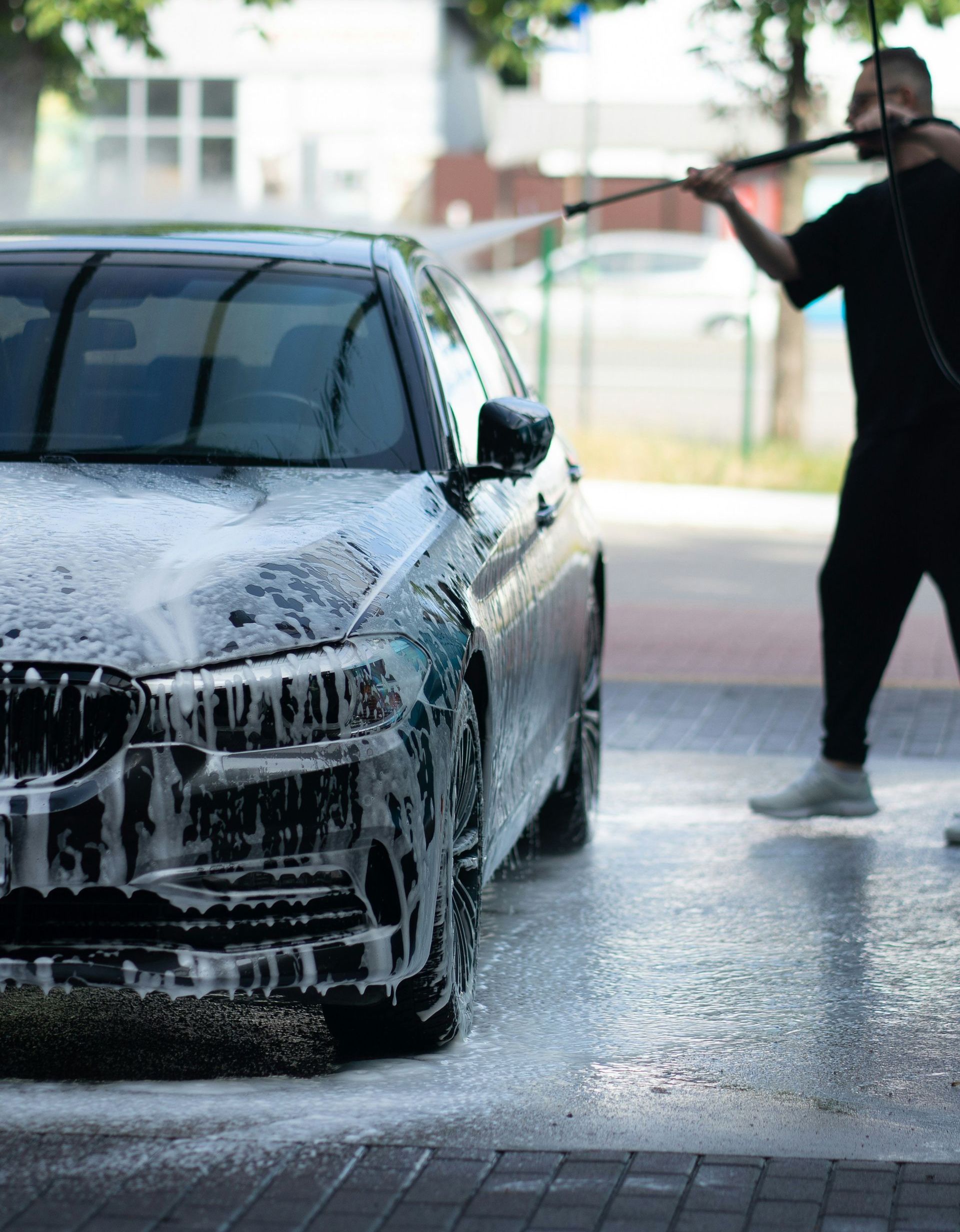 A person in black clothing wipes down a black car in front of a brick and wood building.