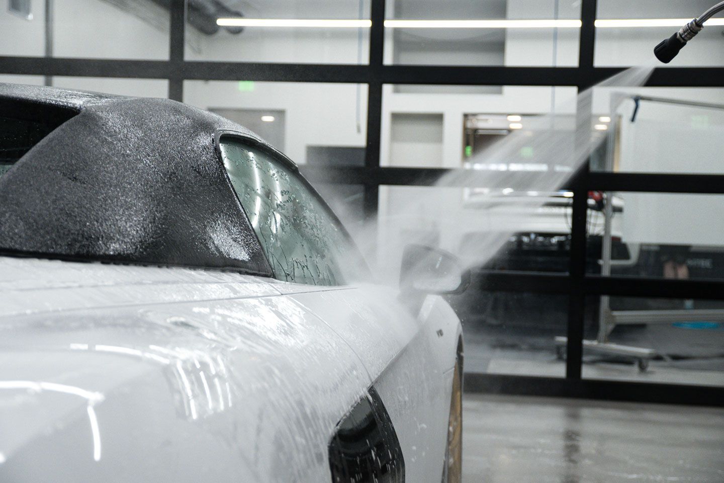 White car being sprayed with water in a car detailing shop. Foam covers the hood and water streams down the side.
