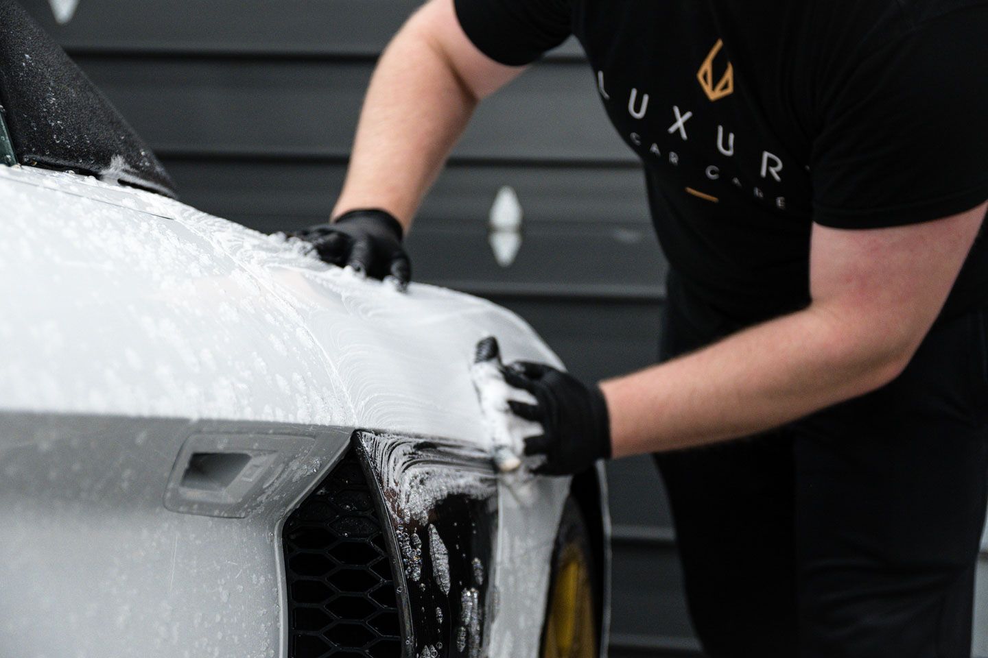 Person washing a white car, covered in soap, with a sponge. They wear black gloves and a black shirt, working outdoors.