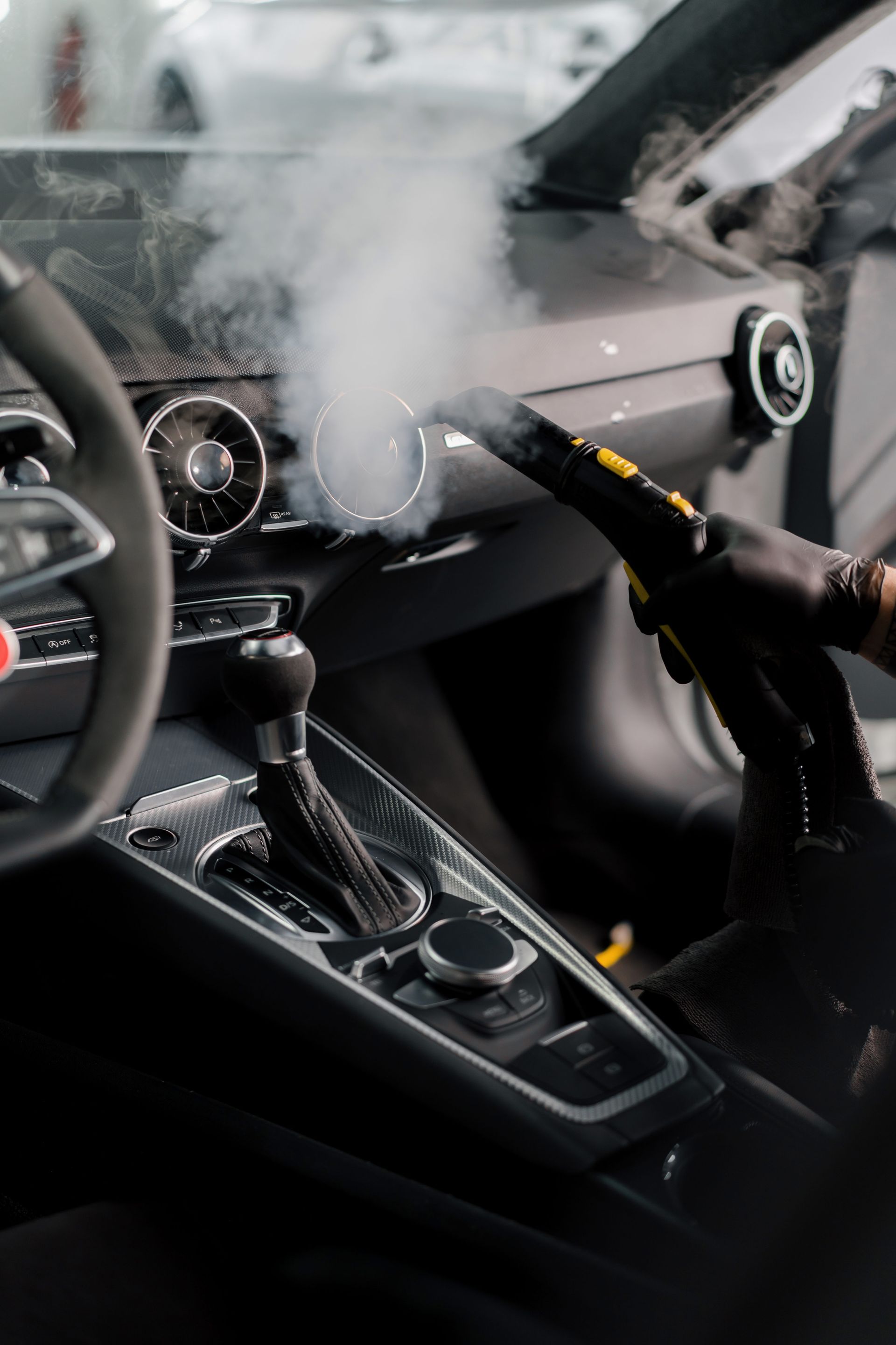 A gloved hand uses a steam cleaner to detail a car's dashboard, releasing a cloud of steam.