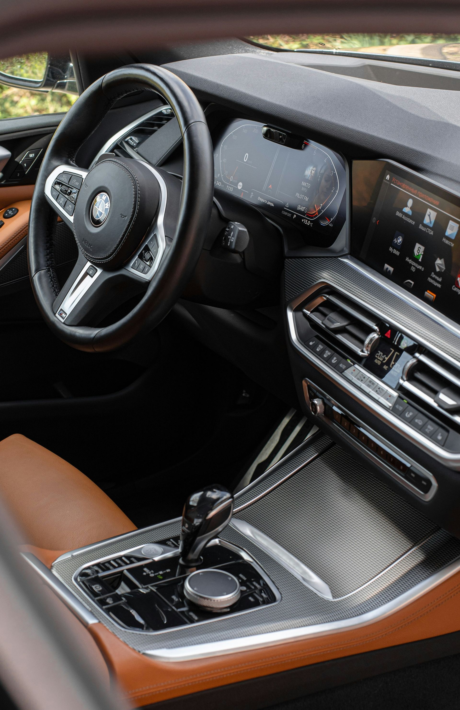 Interior view of a car with a tan leather seat, black steering wheel, and dashboard with a digital display. The console features a carbon fiber-like finish.