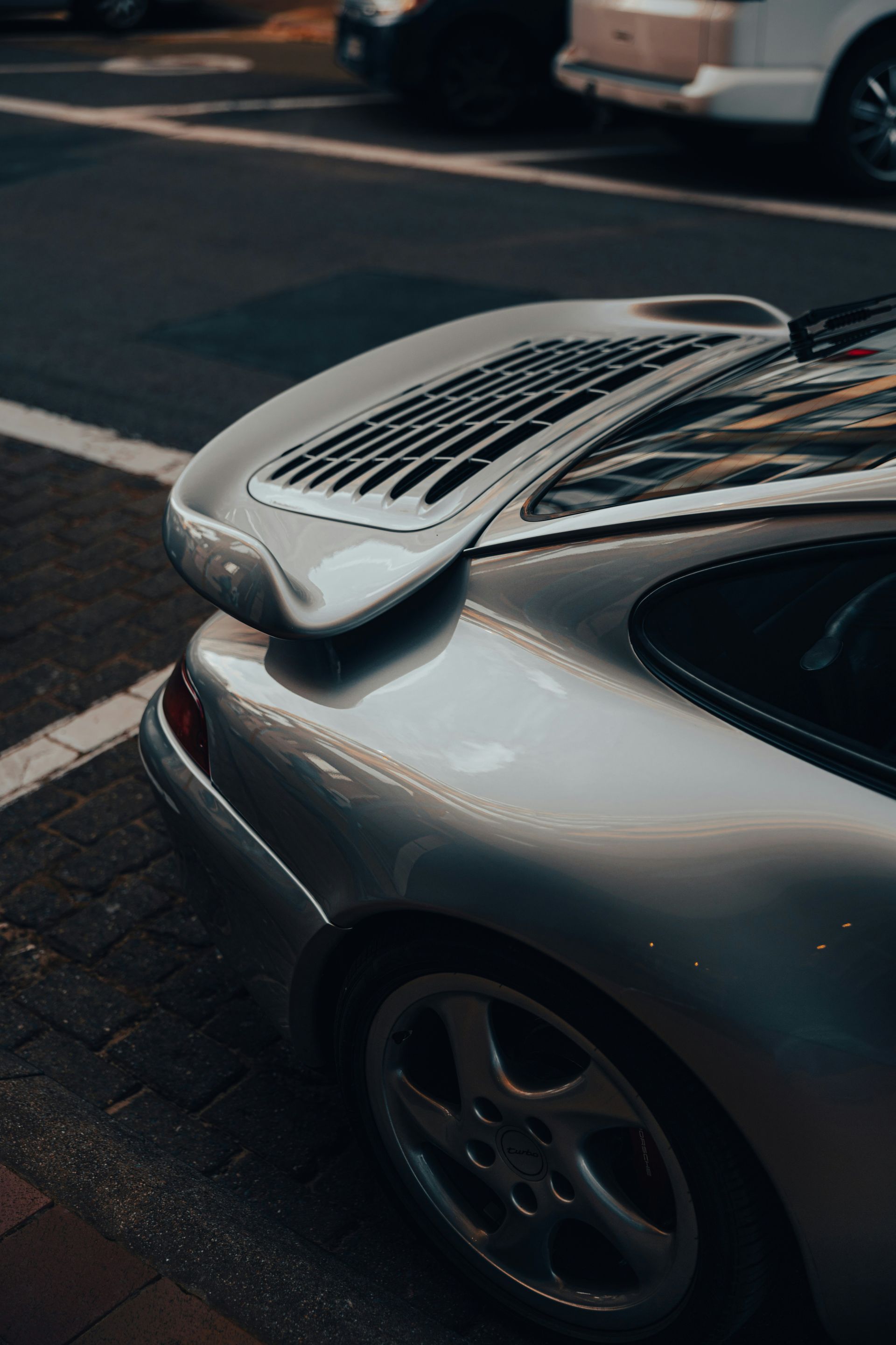Silver Porsche 911 sports car parked on a city street, focusing on the rear and spoiler.