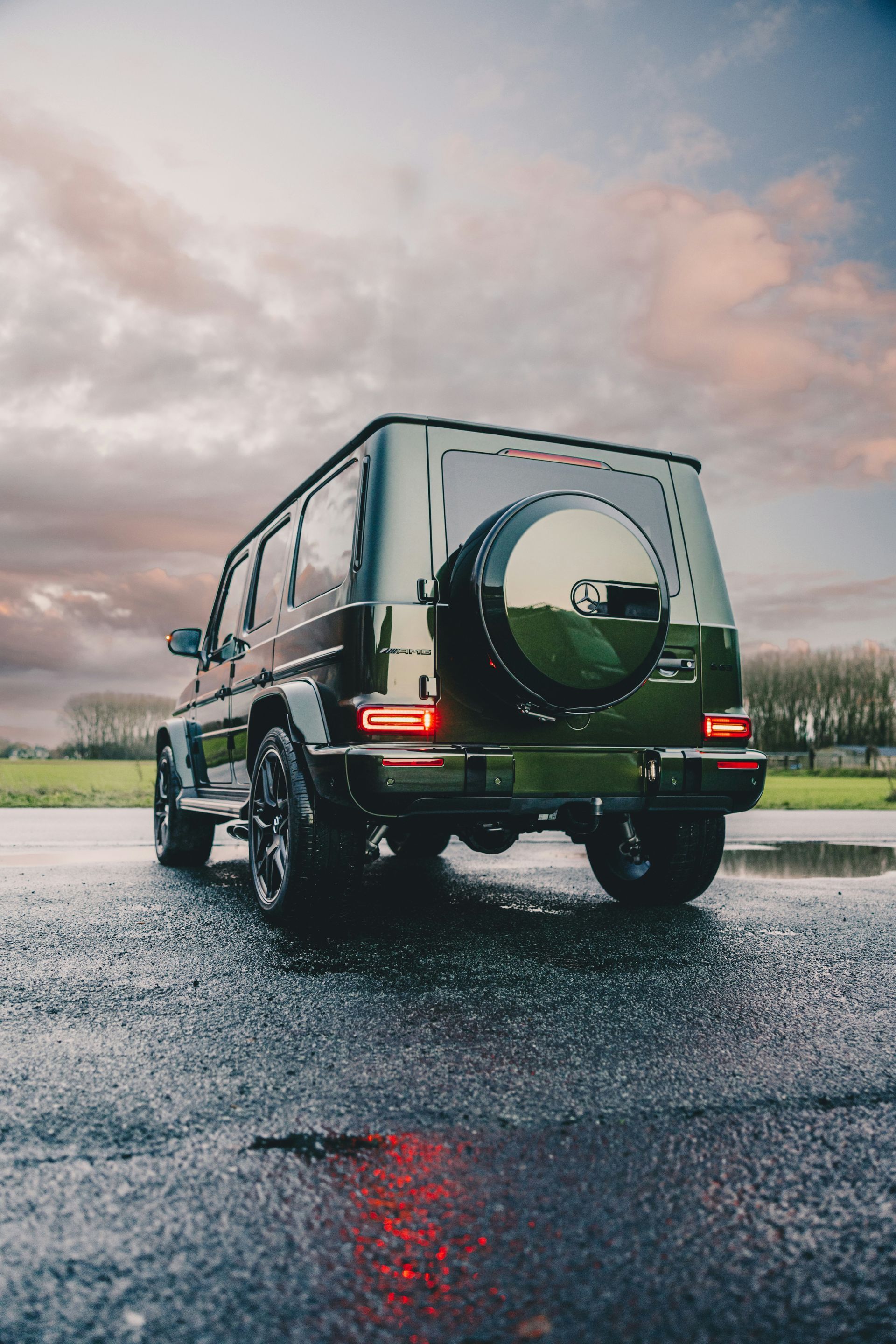 Green Mercedes-Benz G-Class SUV parked on a wet road, with a spare tire on the back and a cloudy sky background.