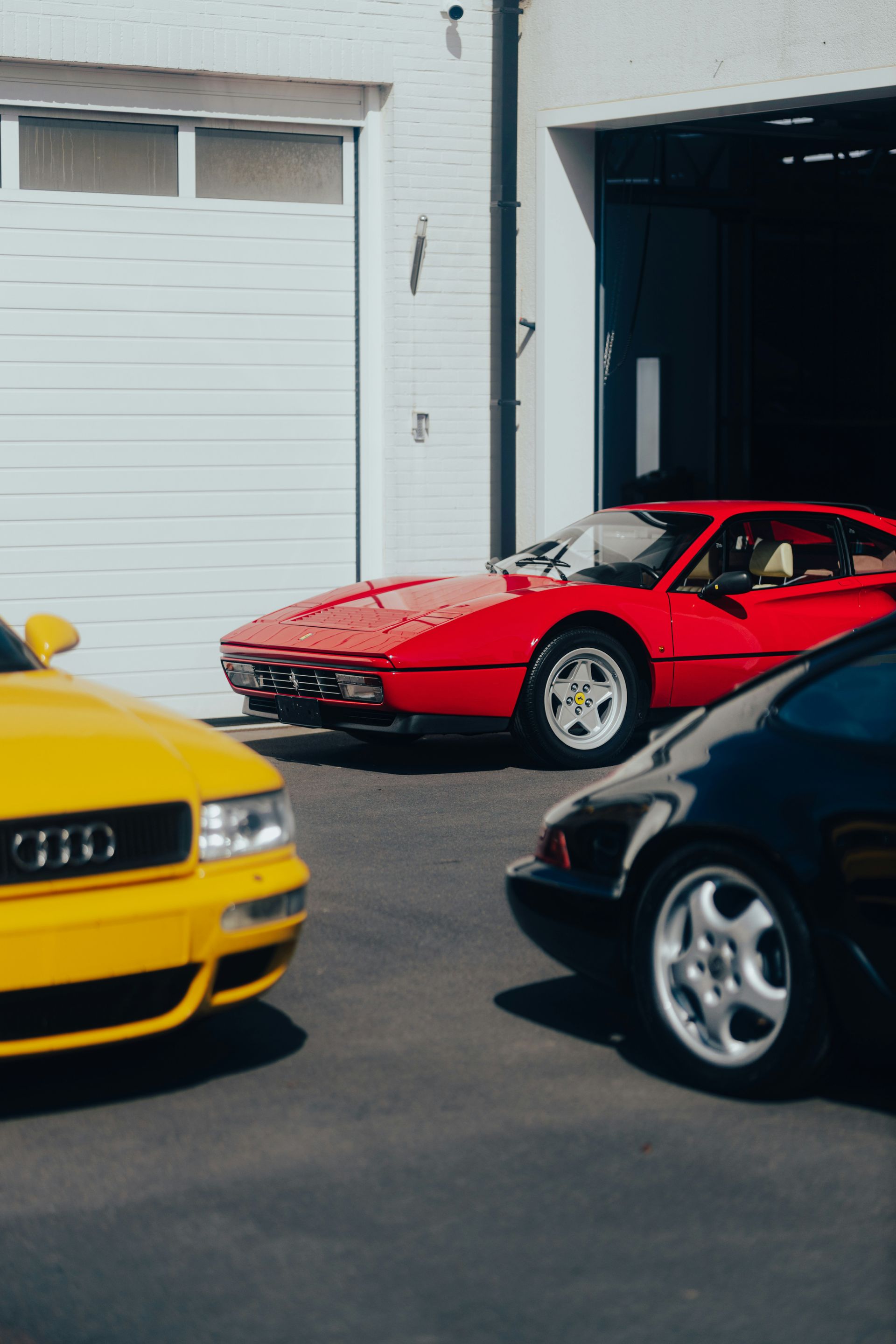Yellow Audi, red Ferrari, and black car parked in front of a garage with a white door.