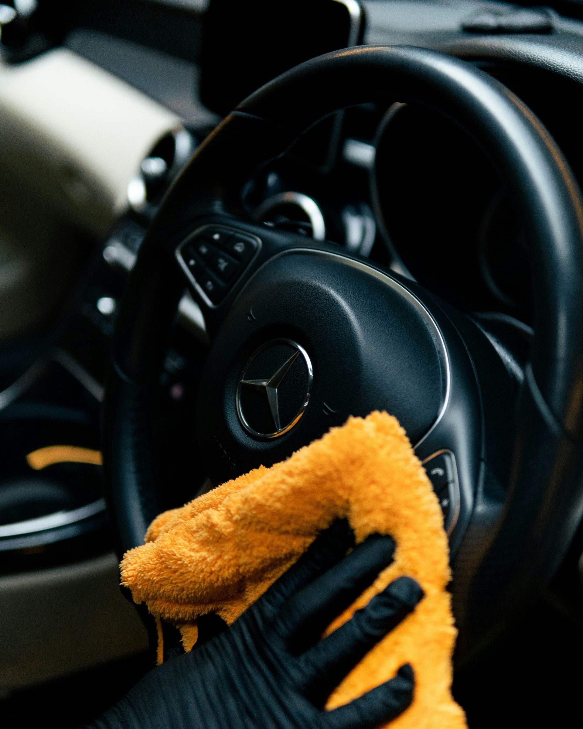 Person wearing black gloves wipes a Mercedes-Benz steering wheel with an orange microfiber cloth.