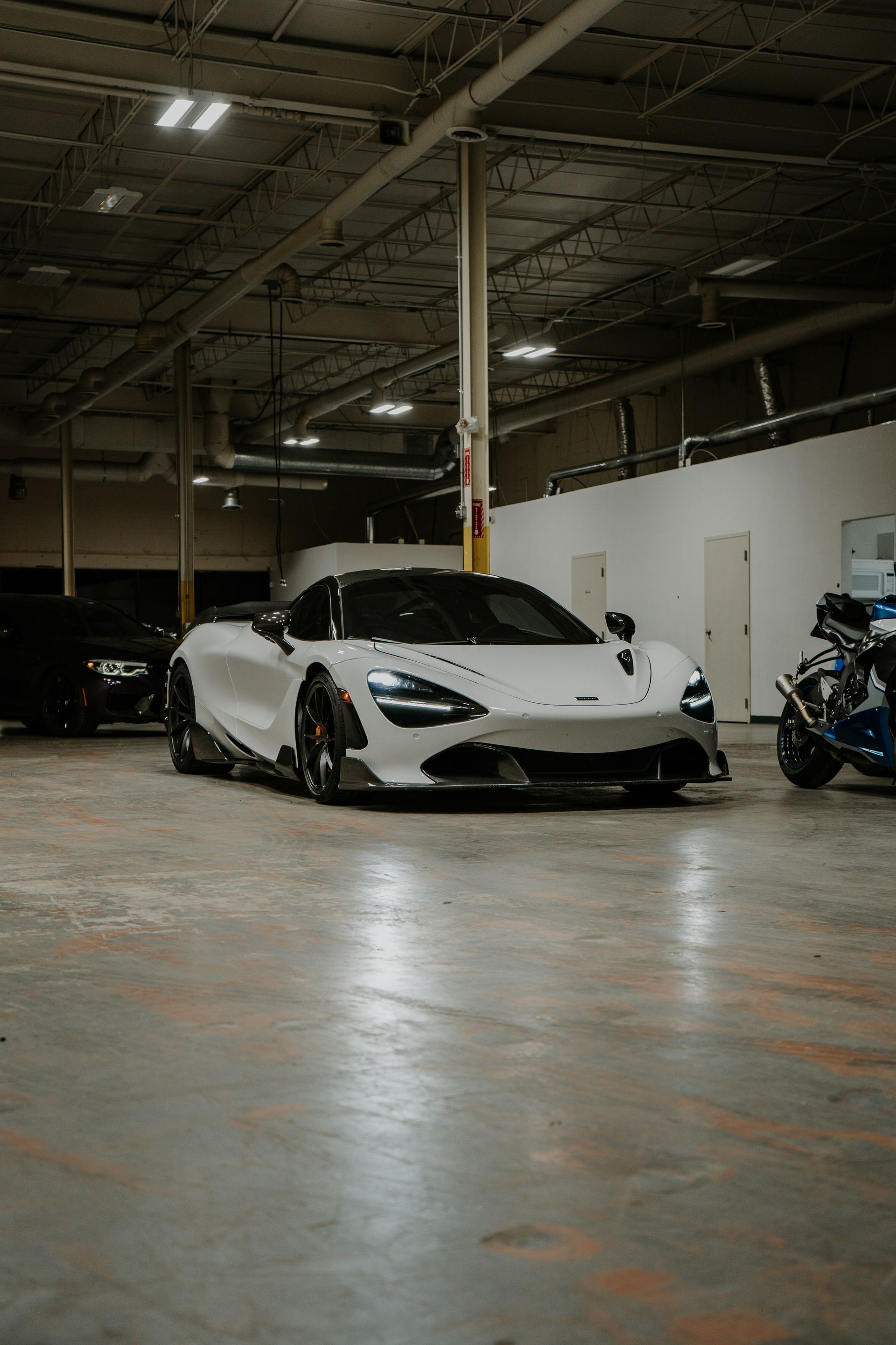 White McLaren sports car parked in a garage with other vehicles. Dimly lit interior with concrete floors.