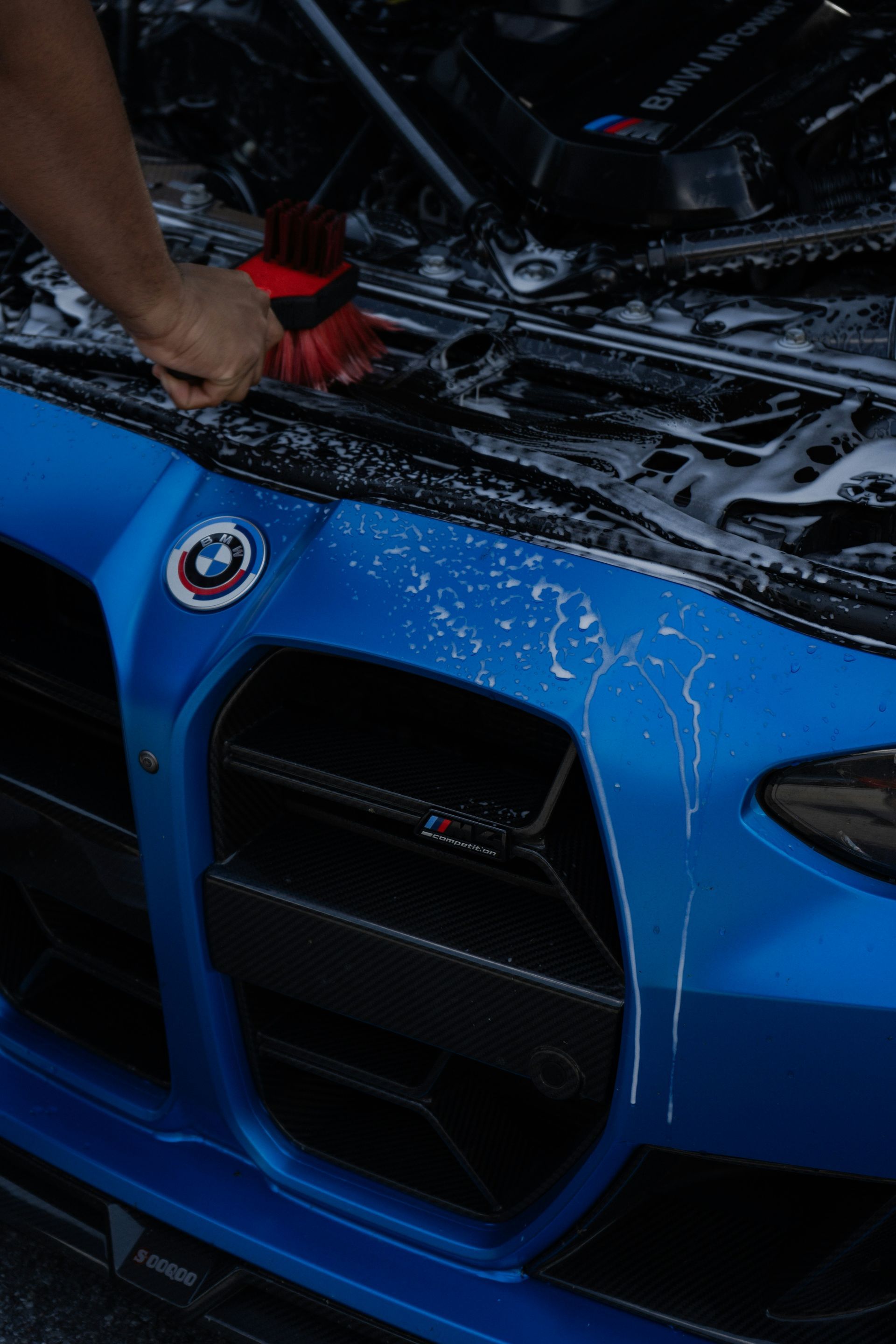 A person cleaning a bright blue BMW car engine bay with a brush and soapy water.