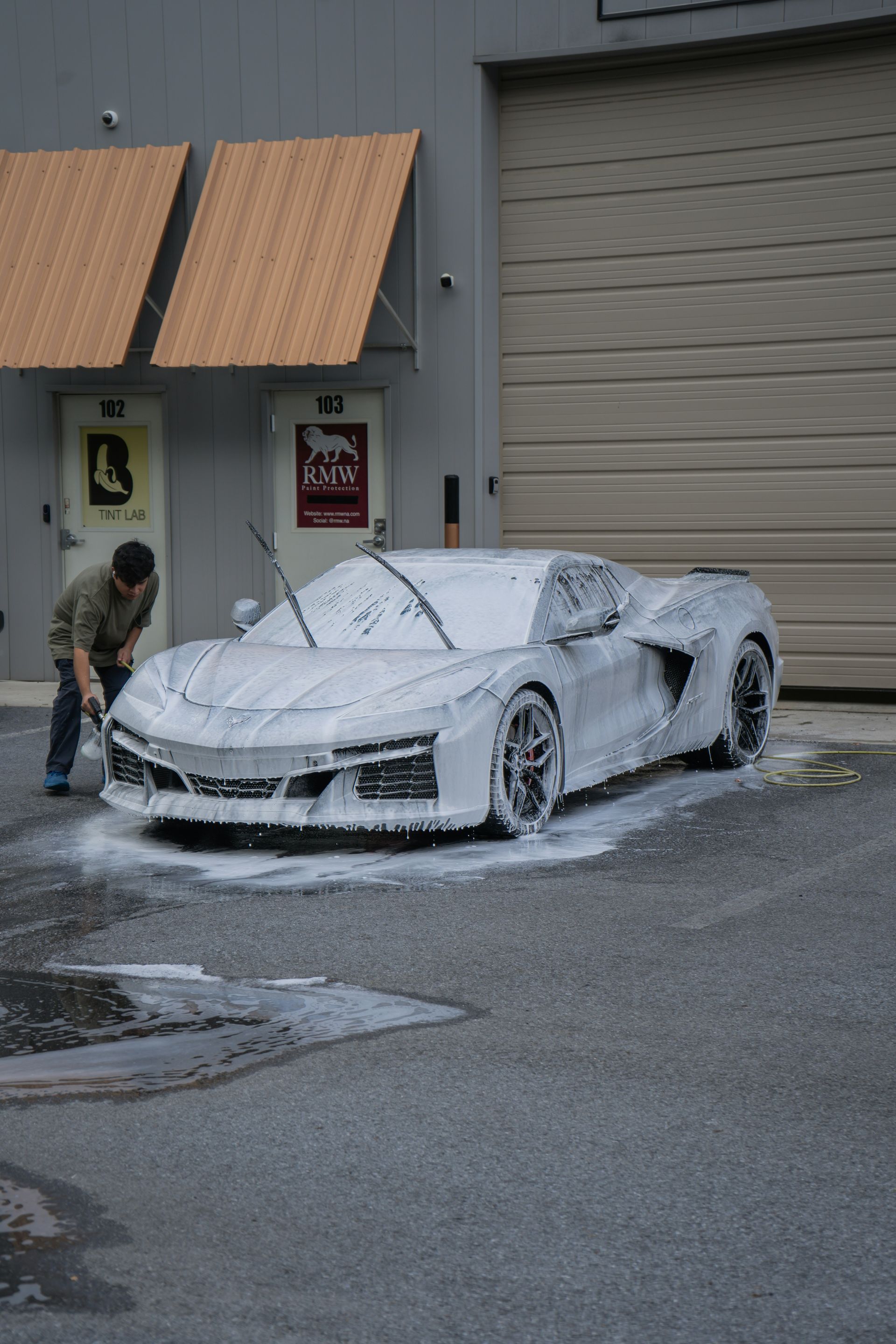 Person washing a gray sports car covered in white foam outside a building with numbered doors.