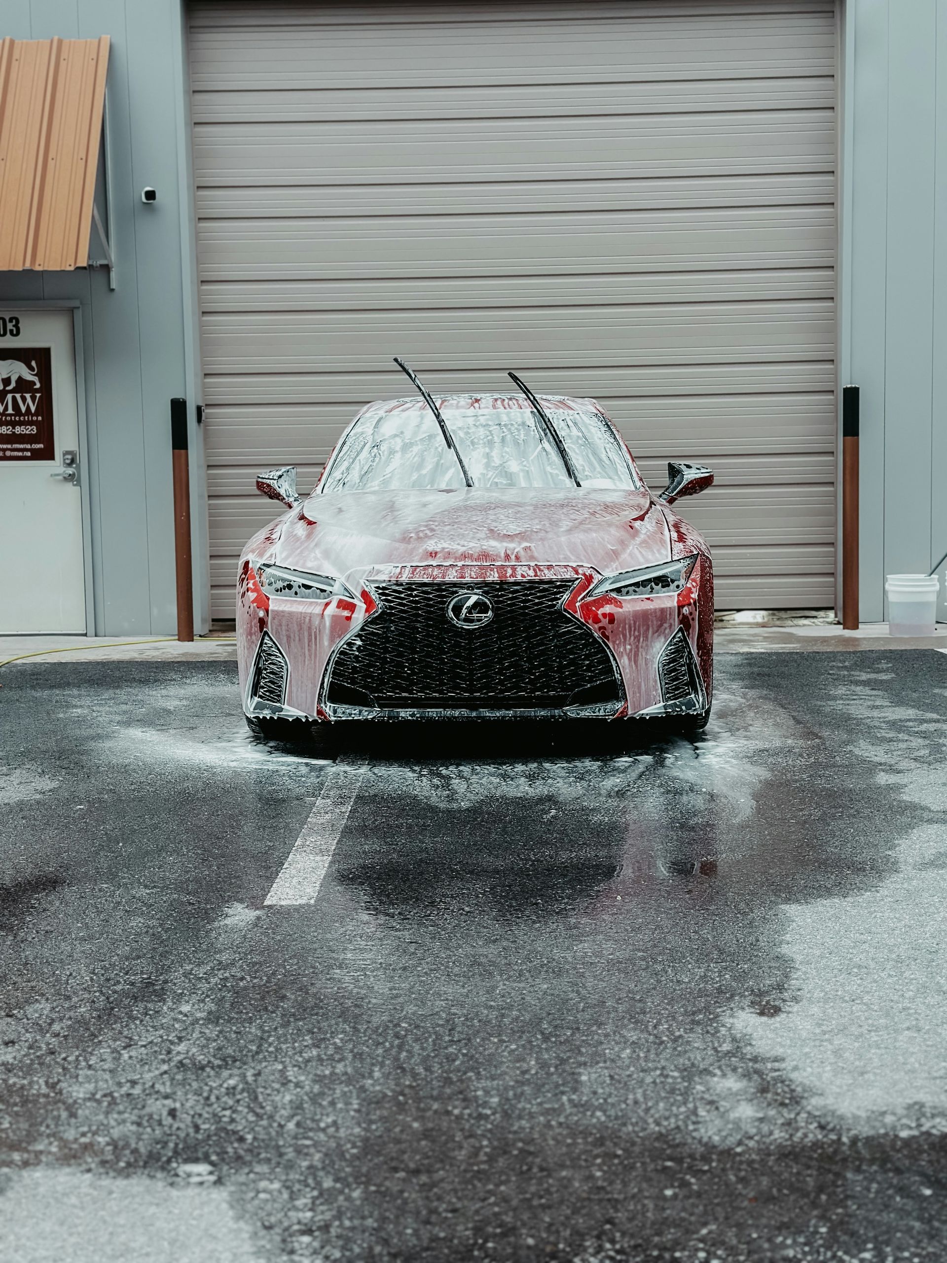 Red Lexus covered in foamy soap, parked in front of a garage door, ready for washing.
