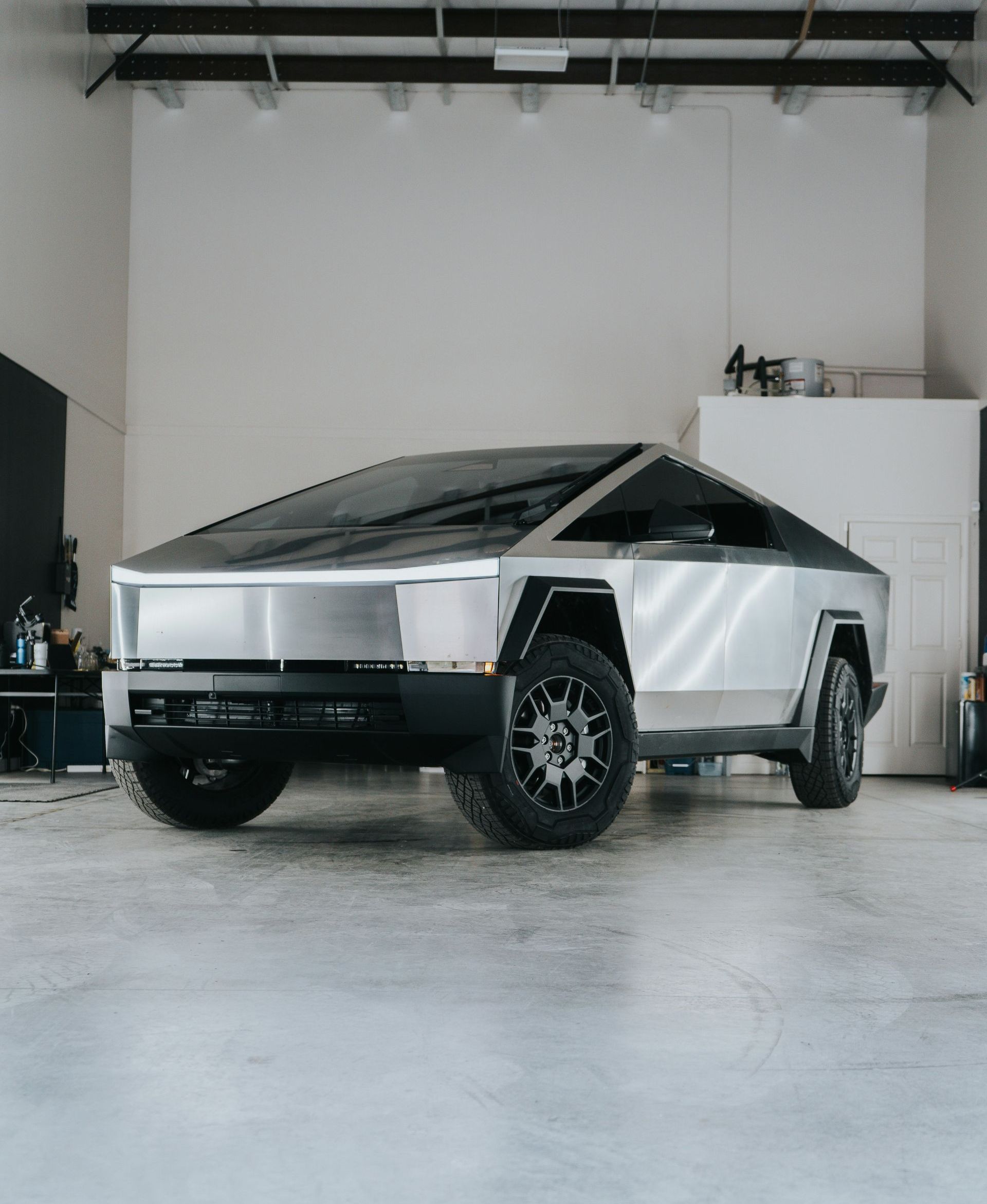 Tesla Cybertruck parked inside a white studio, angled towards the camera. The truck is silver with a futuristic, angular design.