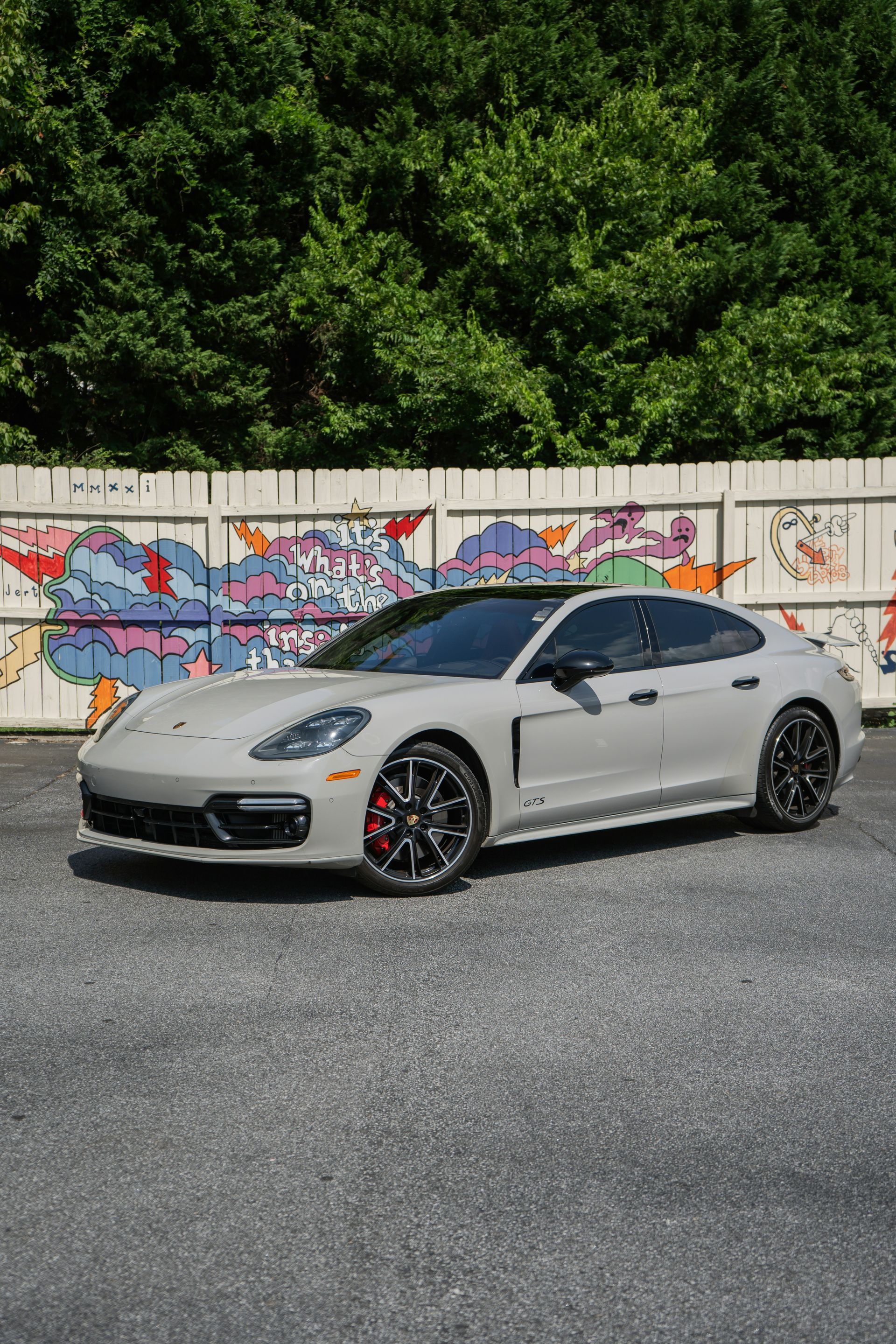 Gray Porsche Panamera parked in front of a wall with colorful graffiti. It has black rims and red brake calipers, set on a gravel surface.