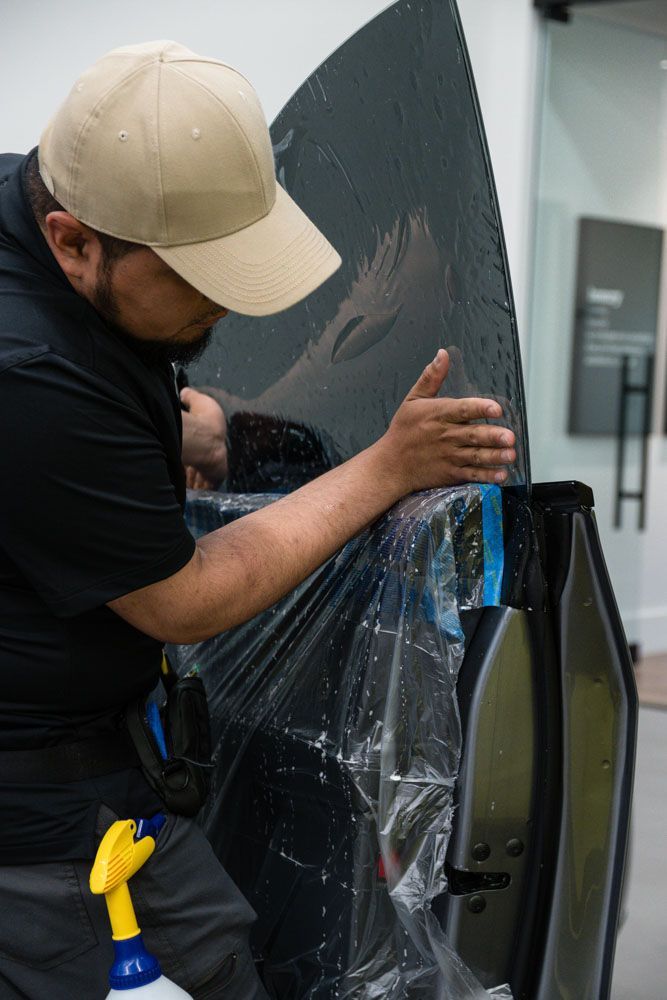 A person in a tan cap applies tinted film to a car window, indoors. They use their hands to smooth the film against the glass.