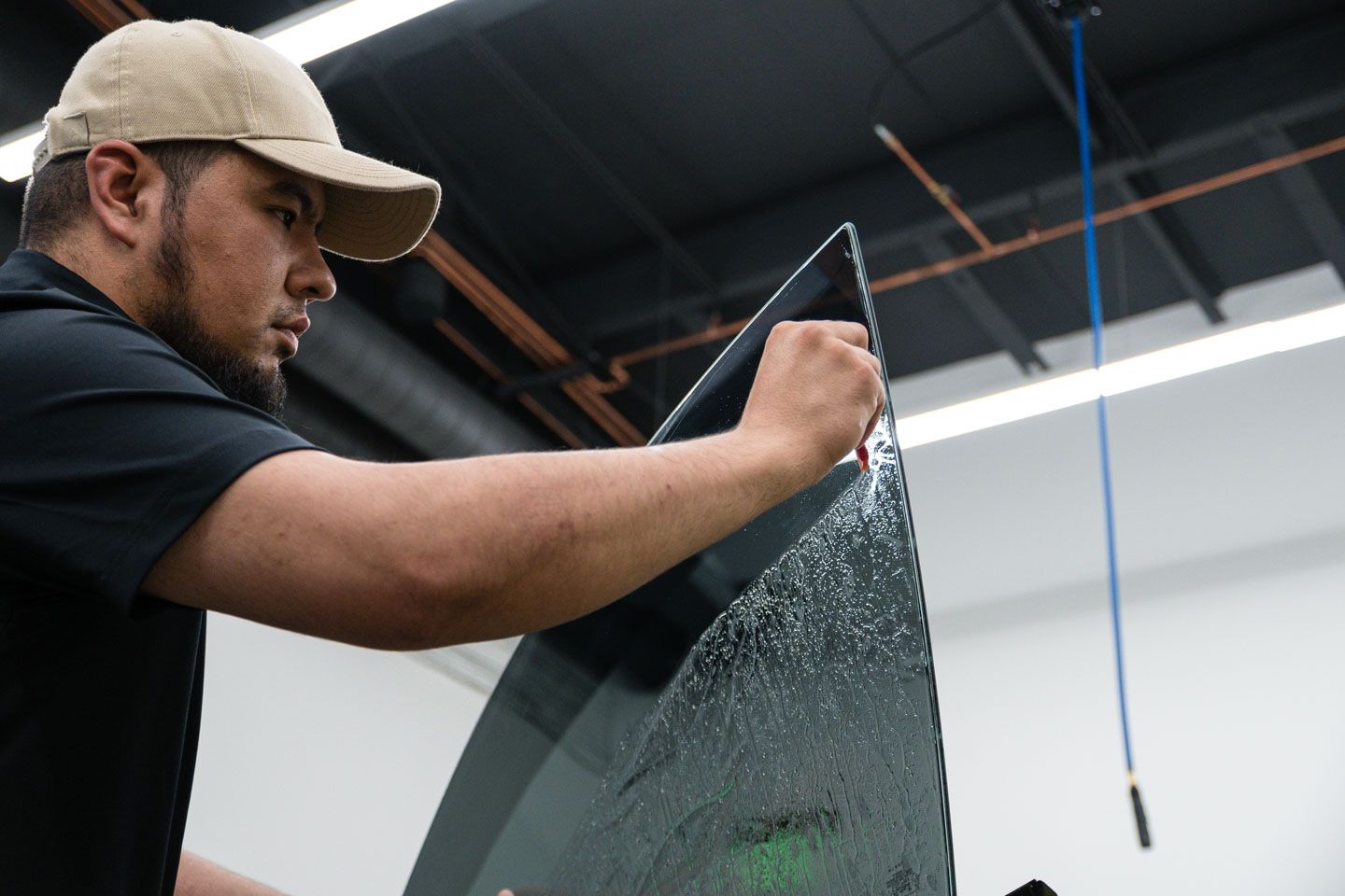 A man in a tan cap is applying window tint to a car window with a squeegee in a bright shop.