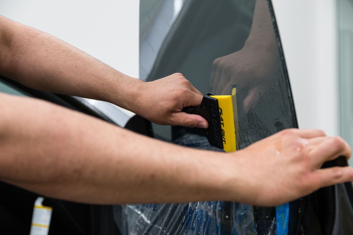 Hands applying window tint to a car window, using a yellow squeegee. The tint is dark, and the setting appears to be an auto shop.