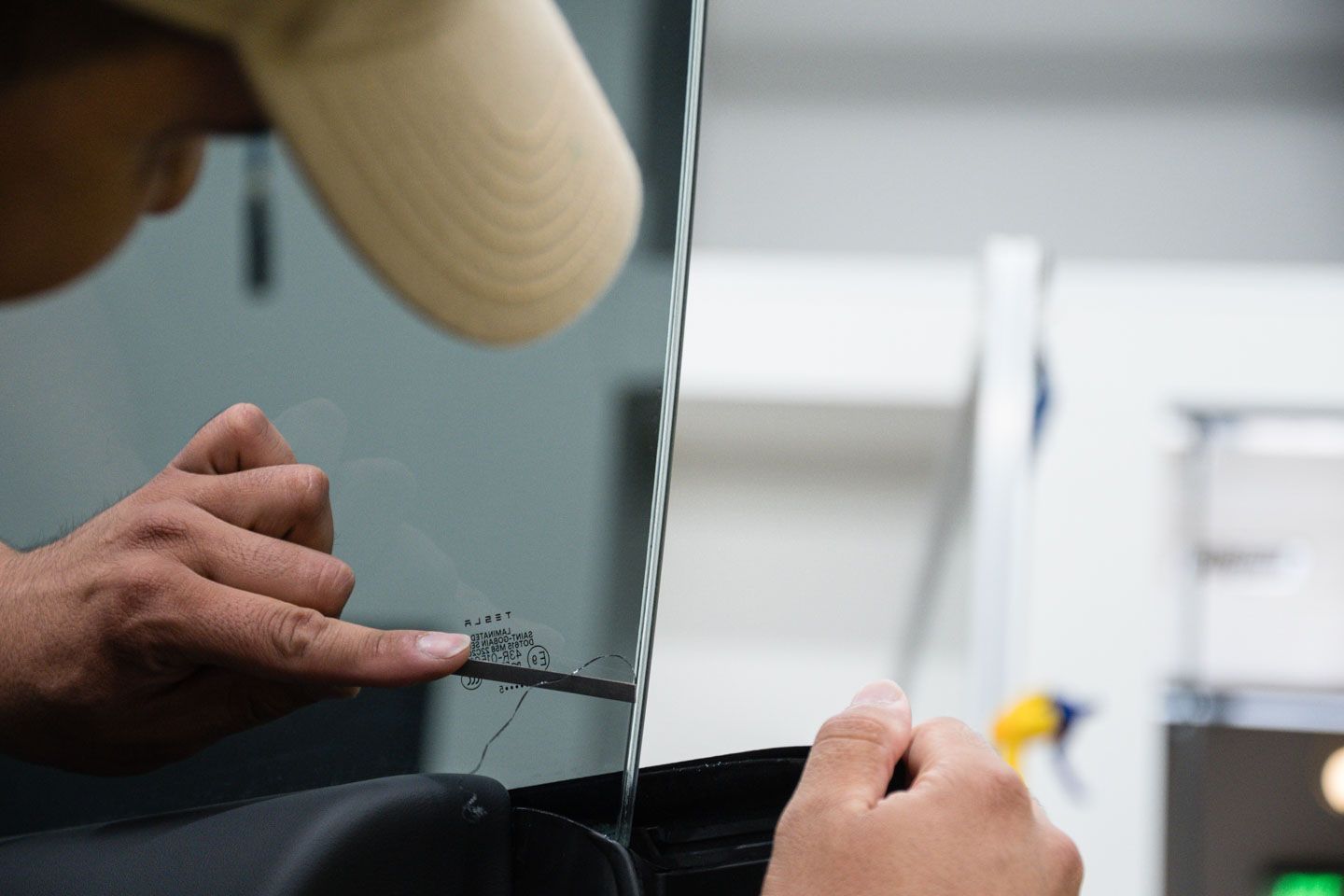 Person applying tint film to a car window, using fingers to smooth it. Wearing a tan cap, inside a garage setting.