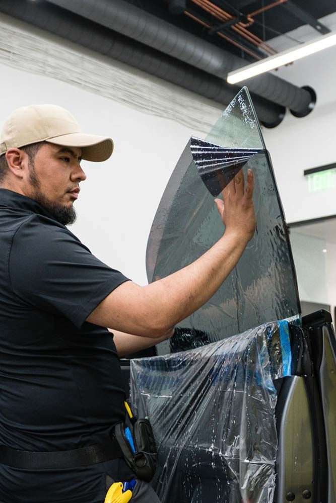 A man wearing a cap, applying tint film to a car window indoors. He is using his hands to smooth the film.