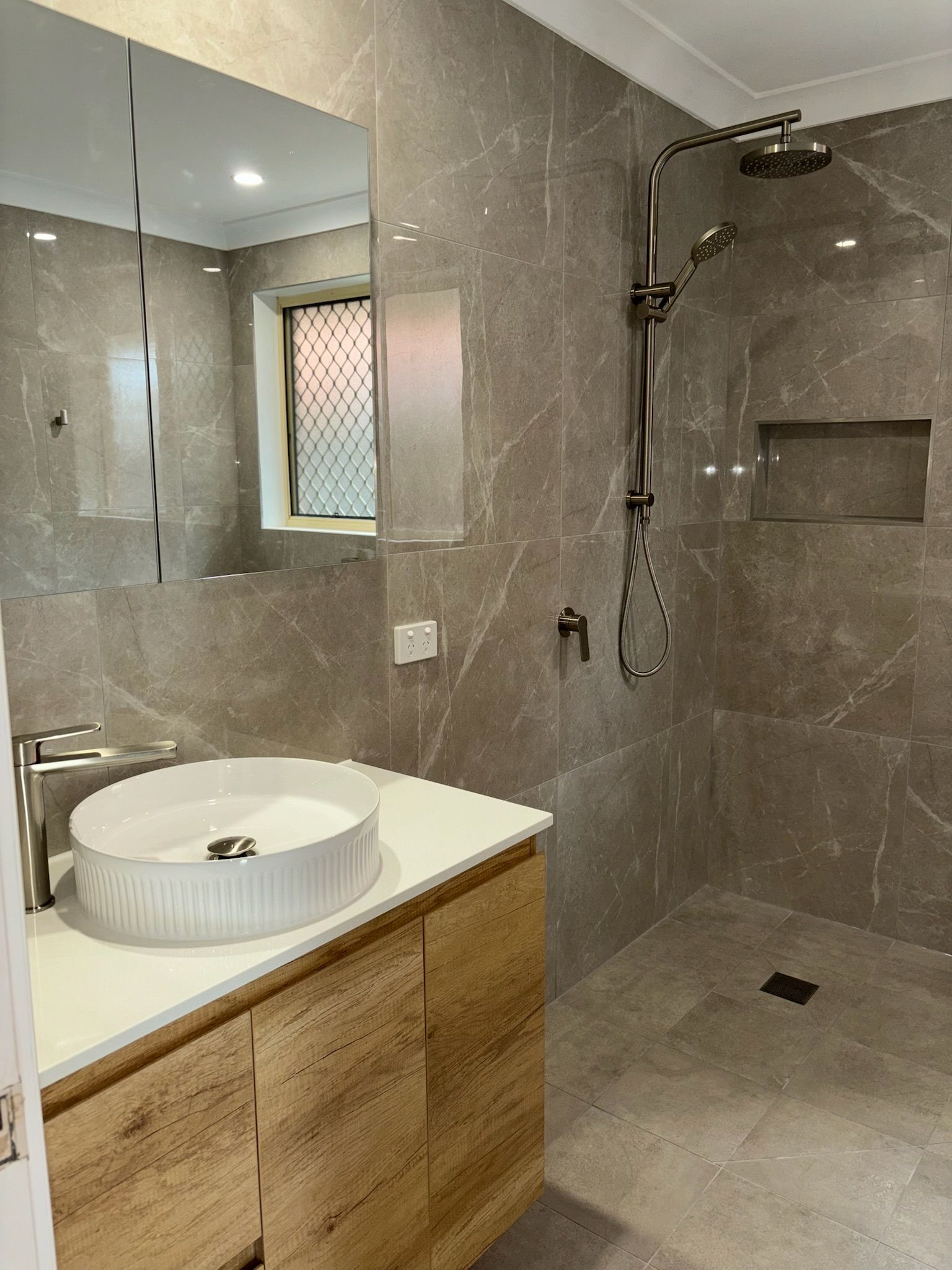 A modern bathroom with a white vessel sink on a wooden vanity, a large wall mirror, and a tiled walk-in shower.