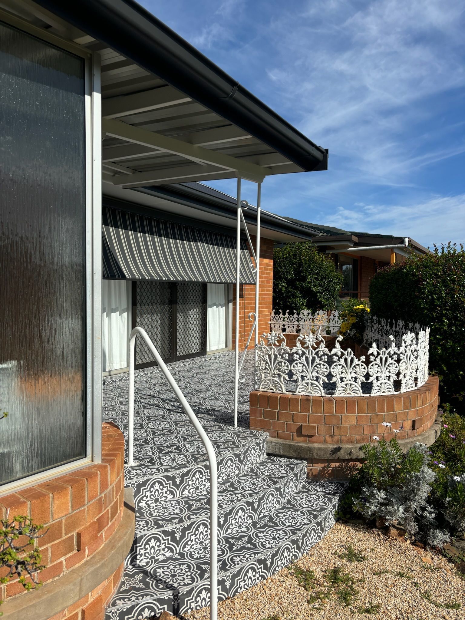 A brick patio and porch area with a patterned black-and-white floor, a white railing, and decorative metal garden fencing.