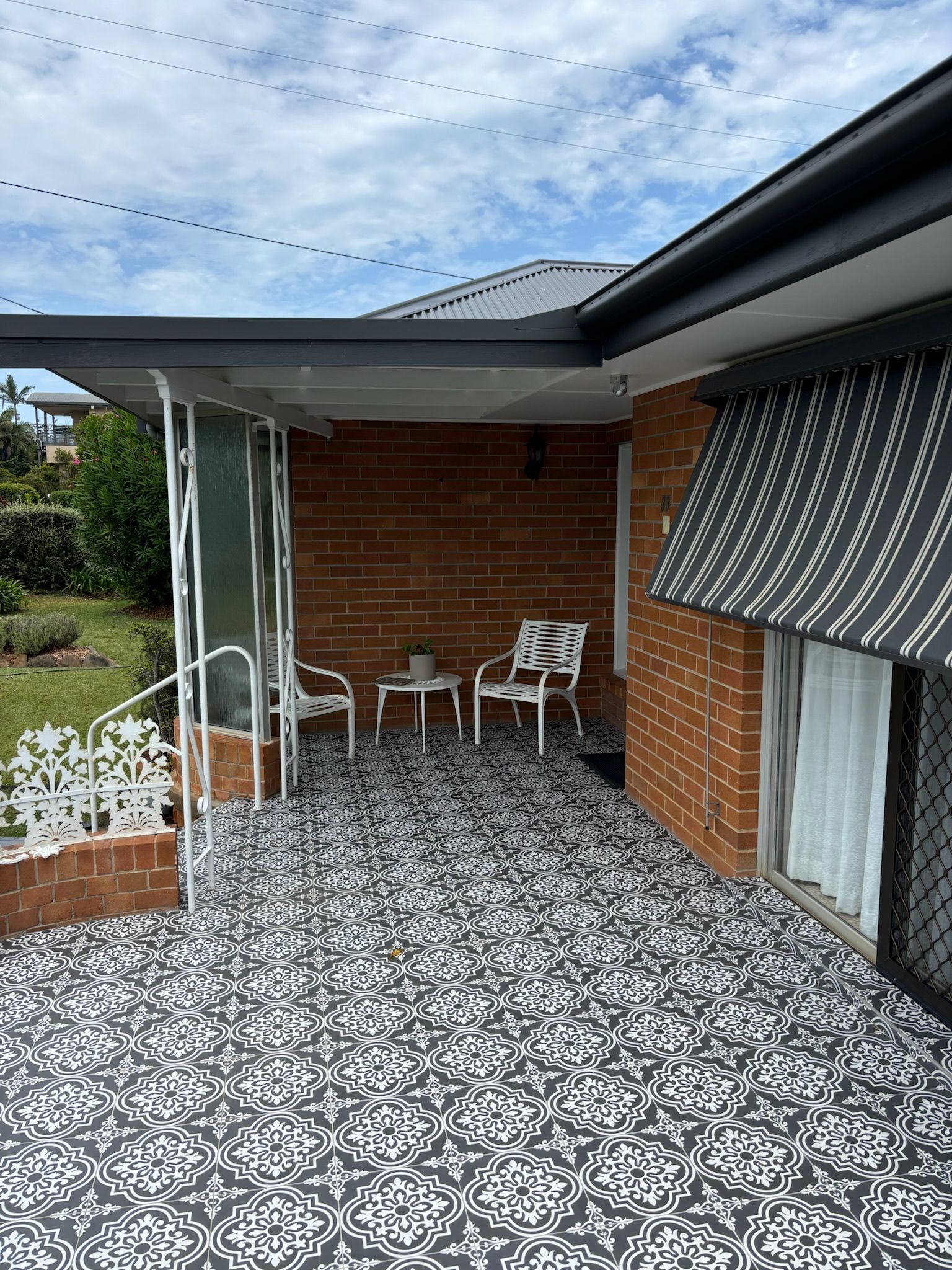 A covered brick patio with a patterned floor, two white chairs, a small table, and a striped awning.
