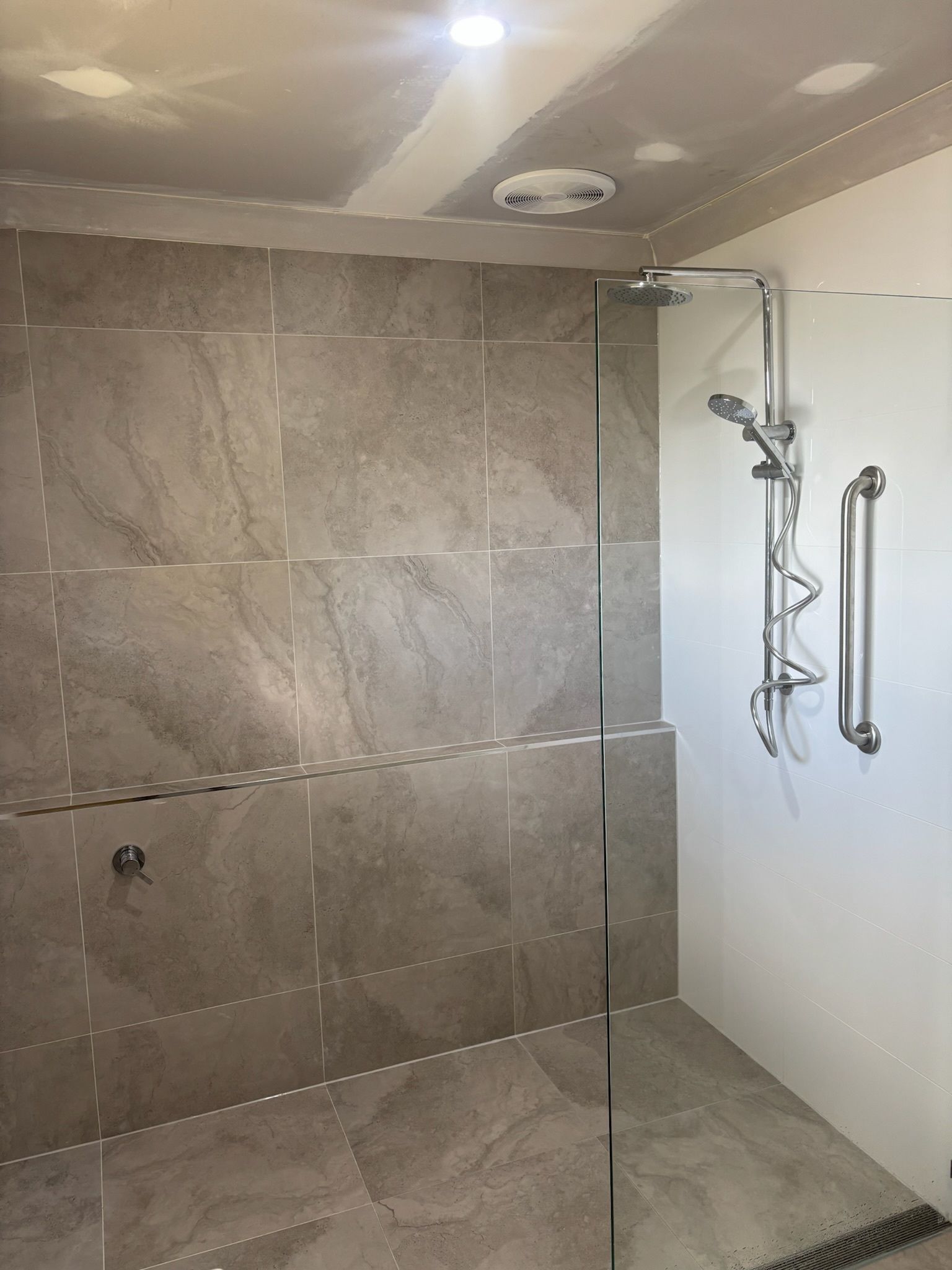 A walk-in shower with grey, stone-look tiled walls, a glass screen, a chrome shower head, and a vertical grab rail.