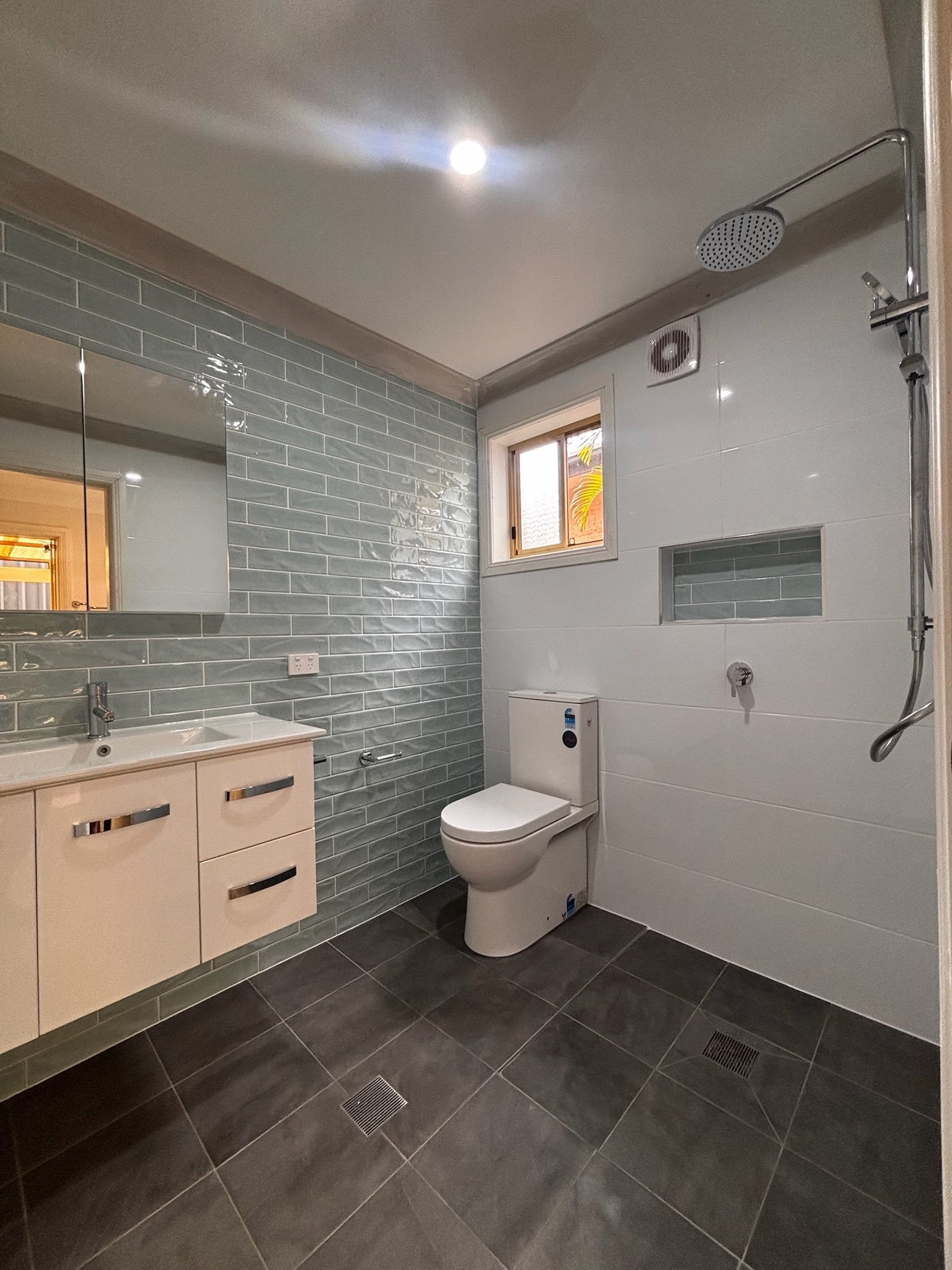 A modern bathroom with grey floor tiles, light blue tiled walls, a wooden vanity, a white toilet, and a walk-in shower.